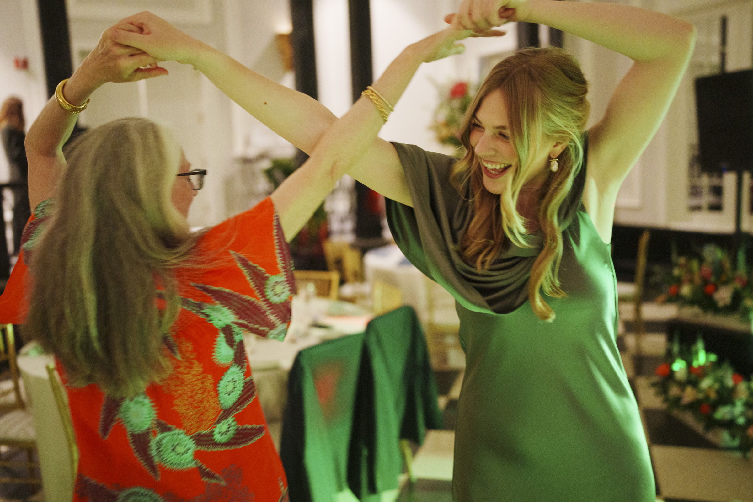 Two women joyfully dance together at an indoor event, smiling and holding hands during an Elegant Spring Celebration. One wears a green dress, the other an orange patterned outfit, perfectly capturing the vibrant spirit of a Carolina Inn Wedding. Tables and decorations are visible in the background. By award-winning Raleigh wedding photographer, Joe Payne.