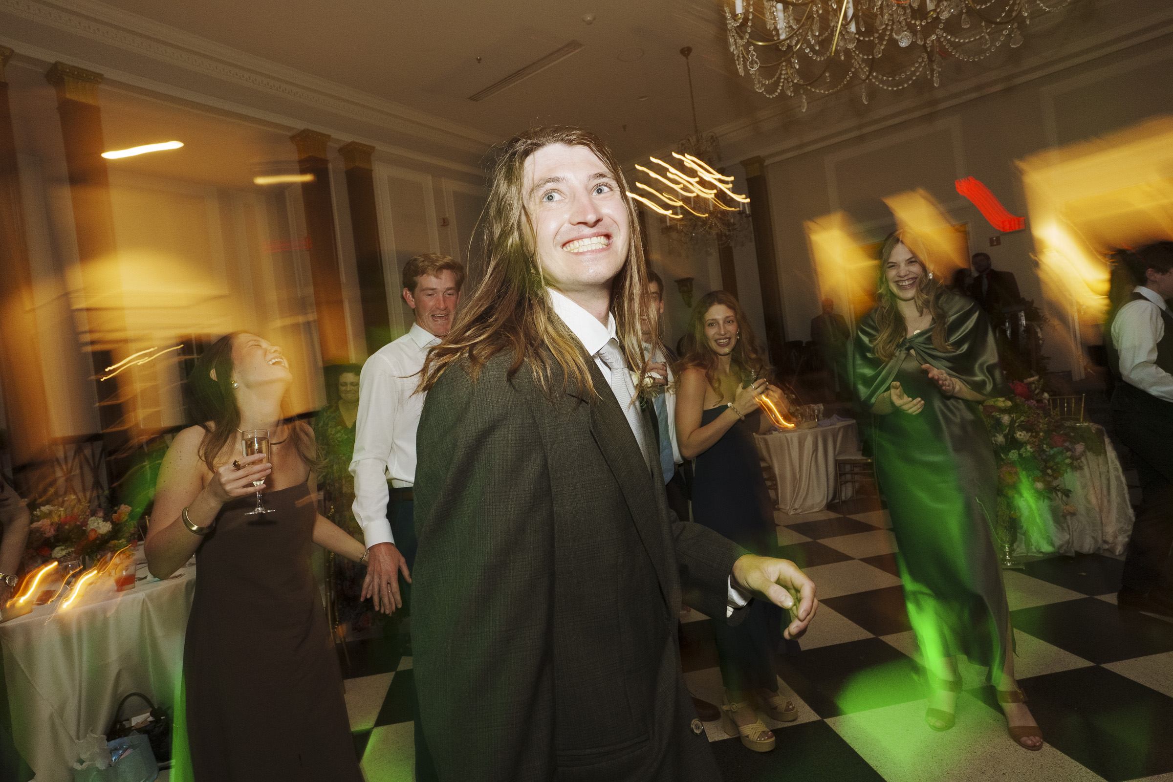 People in formal attire are dancing and smiling at a lively Carolina Inn Wedding in Chapel Hill, with a checkered floor and chandelier. Colorful light effects create a sense of motion and energy throughout this elegant spring celebration. By award-winning Raleigh wedding photographer, Joe Payne.