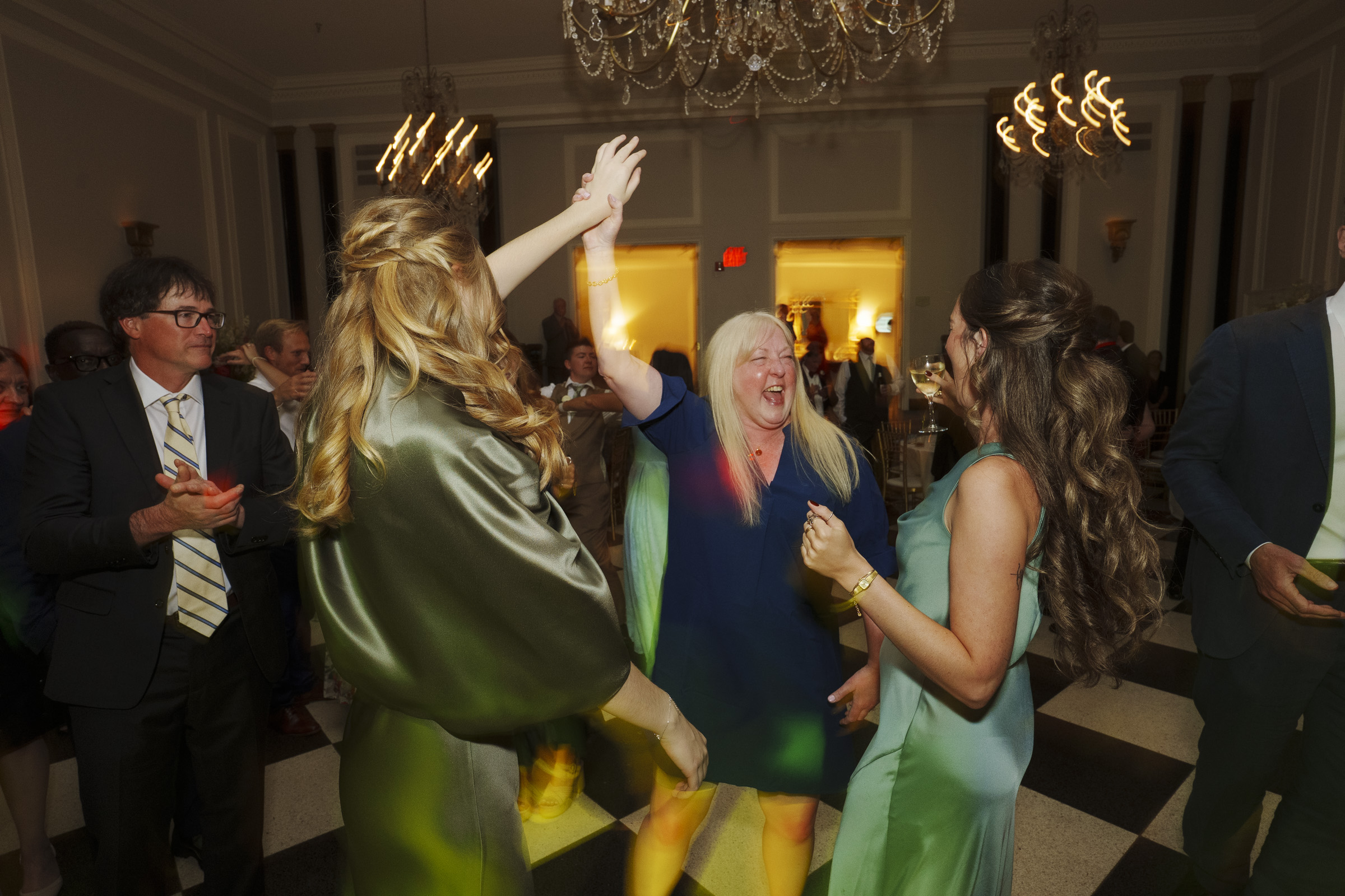 Three women in dresses laugh and dance together at a well-lit indoor event, surrounded by people in formal attire. The atmosphere is lively and joyful, with chandeliers and checkered flooring visible—capturing the vibrant energy of an Elegant Spring Wedding at the Carolina Inn. By award-winning Raleigh wedding photographer, Joe Payne.