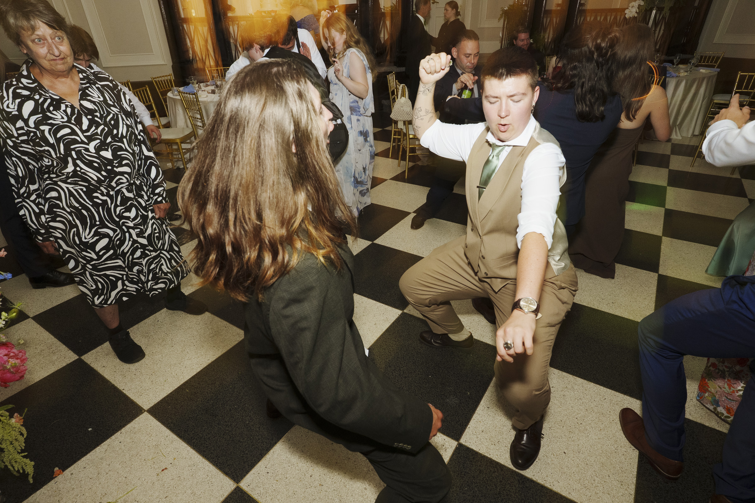 Two people dance energetically at a formal event during an elegant spring celebration at the Carolina Inn Wedding in Chapel Hill, surrounded by other guests. One person in a suit and another in a vest appear to be enjoying the music on a checkered floor while others watch and mingle in the background. By award-winning Raleigh wedding photographer, Joe Payne.