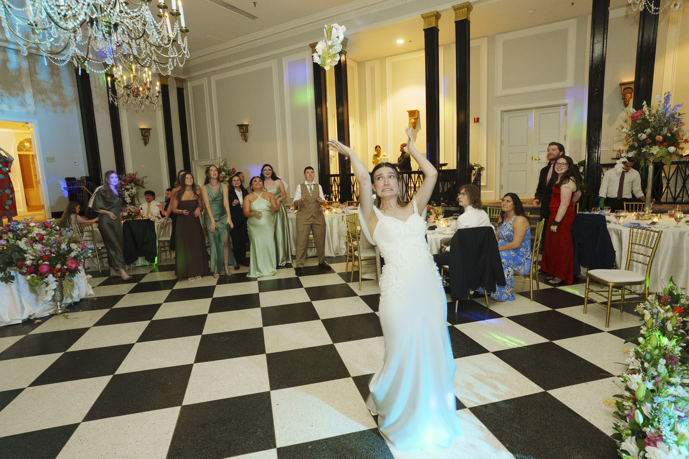A bride in a white gown tosses her bouquet in a ballroom during a Carolina Inn Wedding. Several women behind her, some reaching up, prepare to catch it as part of this Elegant Spring Celebration in Chapel Hill. Guests seated at elegant tables watch as the festivities unfold on the black and white checkered floor. By award-winning Raleigh wedding photographer, Joe Payne.
