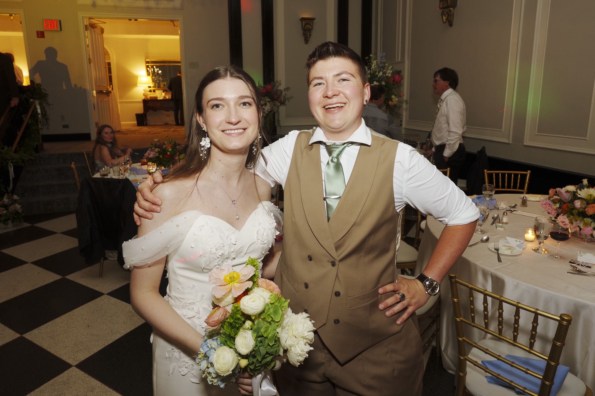 Two people smile together at a formal event at the Carolina Inn in Chapel Hill. One wears a white dress and holds a bouquet; the other wears a tan vest and tie. Elegant tables and floral decorations set the scene for this Elegant Spring Celebration in the background. By award-winning Raleigh wedding photographer, Joe Payne.