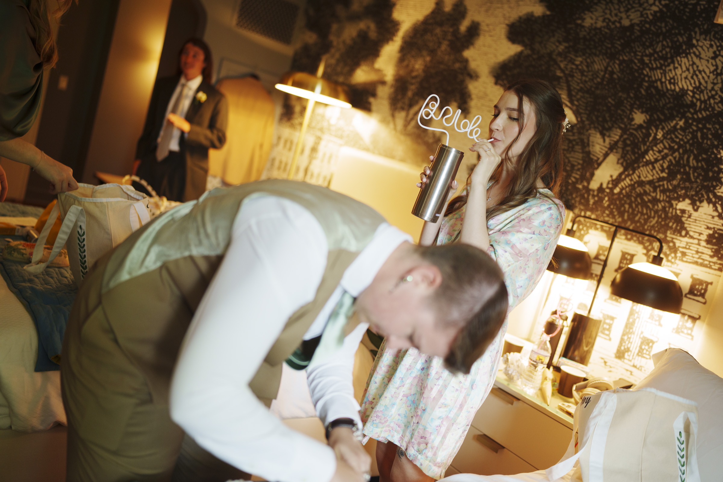 A woman in a floral dress sips from a cup with a curly straw labeled “bride” while a man in a tan suit bends over nearby in a warmly lit hotel room, capturing the joyful atmosphere of an Elegant Spring Celebration. Another man in a suit stands in the background, adding to the anticipation and excitement of this beautiful Carolina Inn Wedding in Chapel Hill. By award-winning Raleigh wedding photographer, Joe Payne.