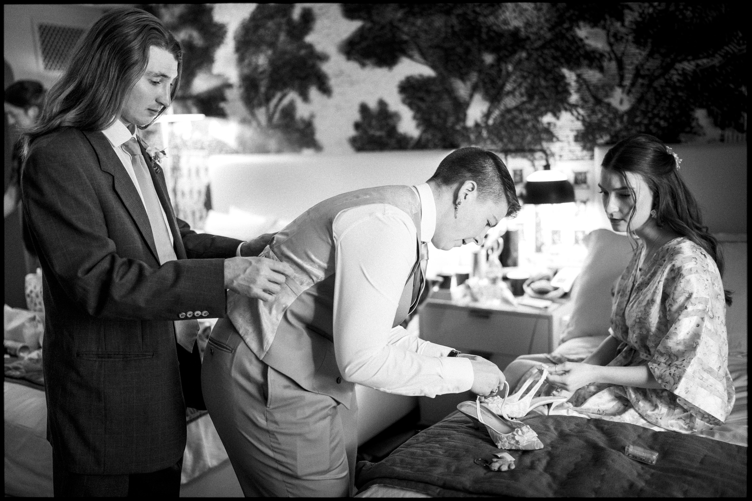 Three people in a bedroom: one person helps another adjust a vest, while a third, seated on the bed in a robe, looks on. The room is softly lit with patterned wallpaper in the background, capturing an intimate moment of preparation before an Elegant Spring Celebration at the Carolina Inn Wedding. By award-winning Raleigh wedding photographer, Joe Payne.