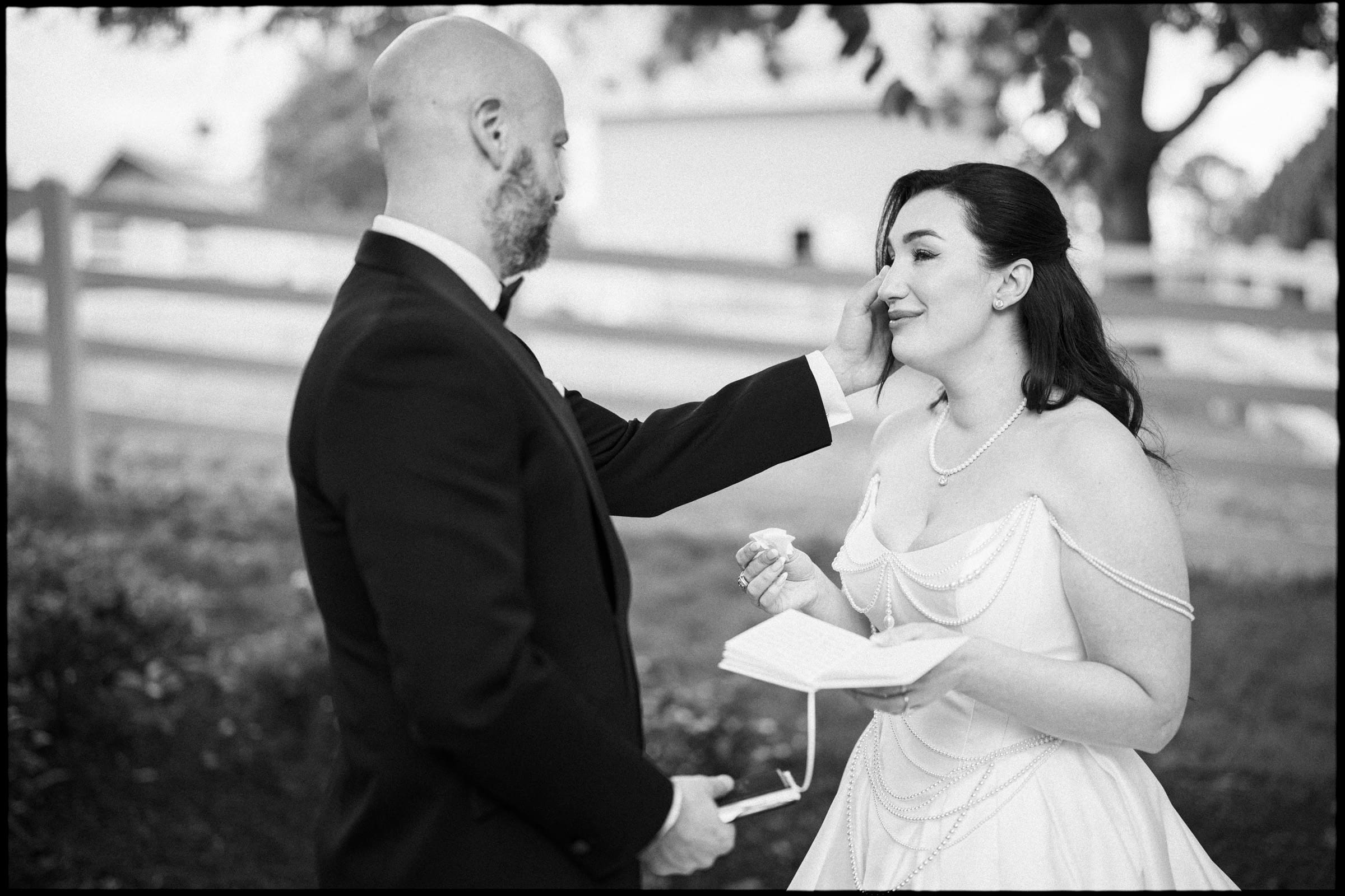 A groom gently touches the bride’s face as she smiles and wipes a tear, holding a small notebook and glasses. At their Historic Wakefield Barn Wedding, they’re outdoors, dressed formally, with a wooden fence and trees in the background. By award-winning Raleigh wedding photographer, Joe Payne.