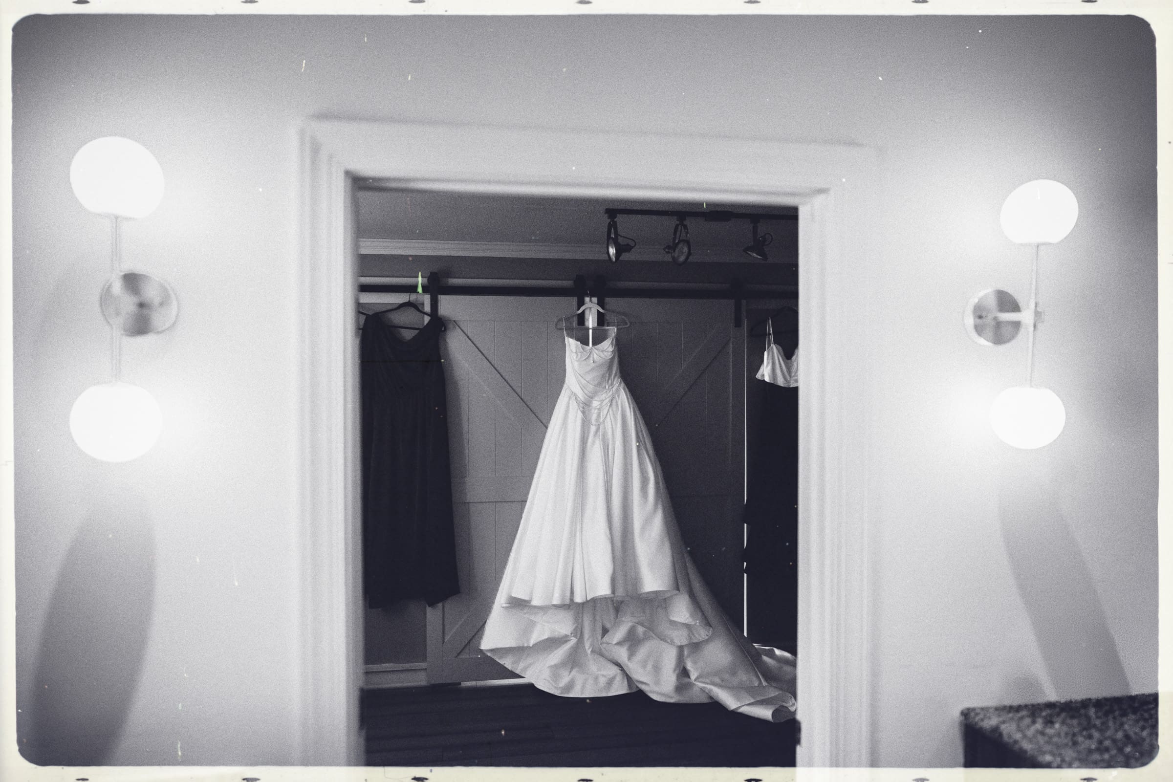 A white wedding dress hangs on a hanger in the center of a doorway, flanked by two glowing wall lamps. The black and white photo captures the nostalgic, timeless feel of a Wakefield barn wedding. By award-winning Raleigh wedding photographer, Joe Payne.
