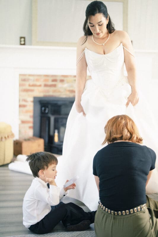 A bride in a white gown stands as a woman kneels to adjust her dress. A young boy in a dress shirt and bow tie sits nearby, looking at a piece of paper. The softly lit room with a fireplace sets the scene for this Wakefield Barn wedding. By award-winning Raleigh wedding photographer, Joe Payne.