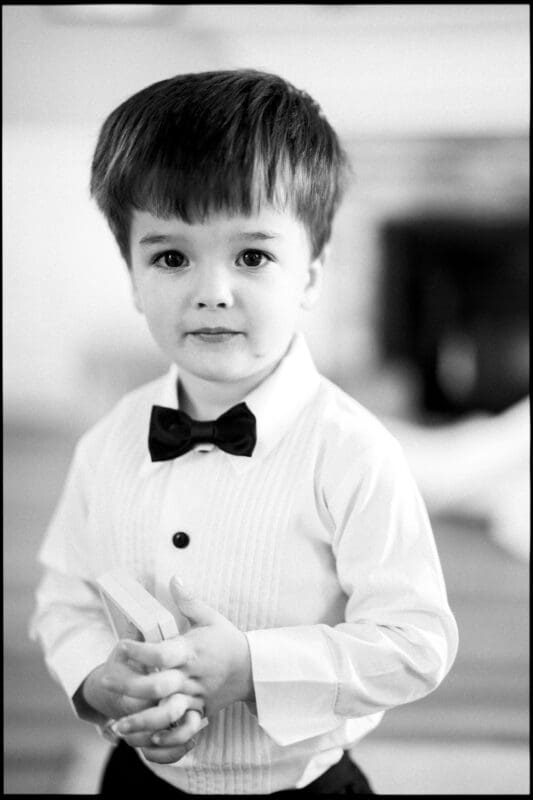 A young boy dressed in a white dress shirt with a black bow tie stands indoors at a Wakefield Barn wedding, looking at the camera while holding something in his hands. The image is in black and white. By award-winning Raleigh wedding photographer, Joe Payne.