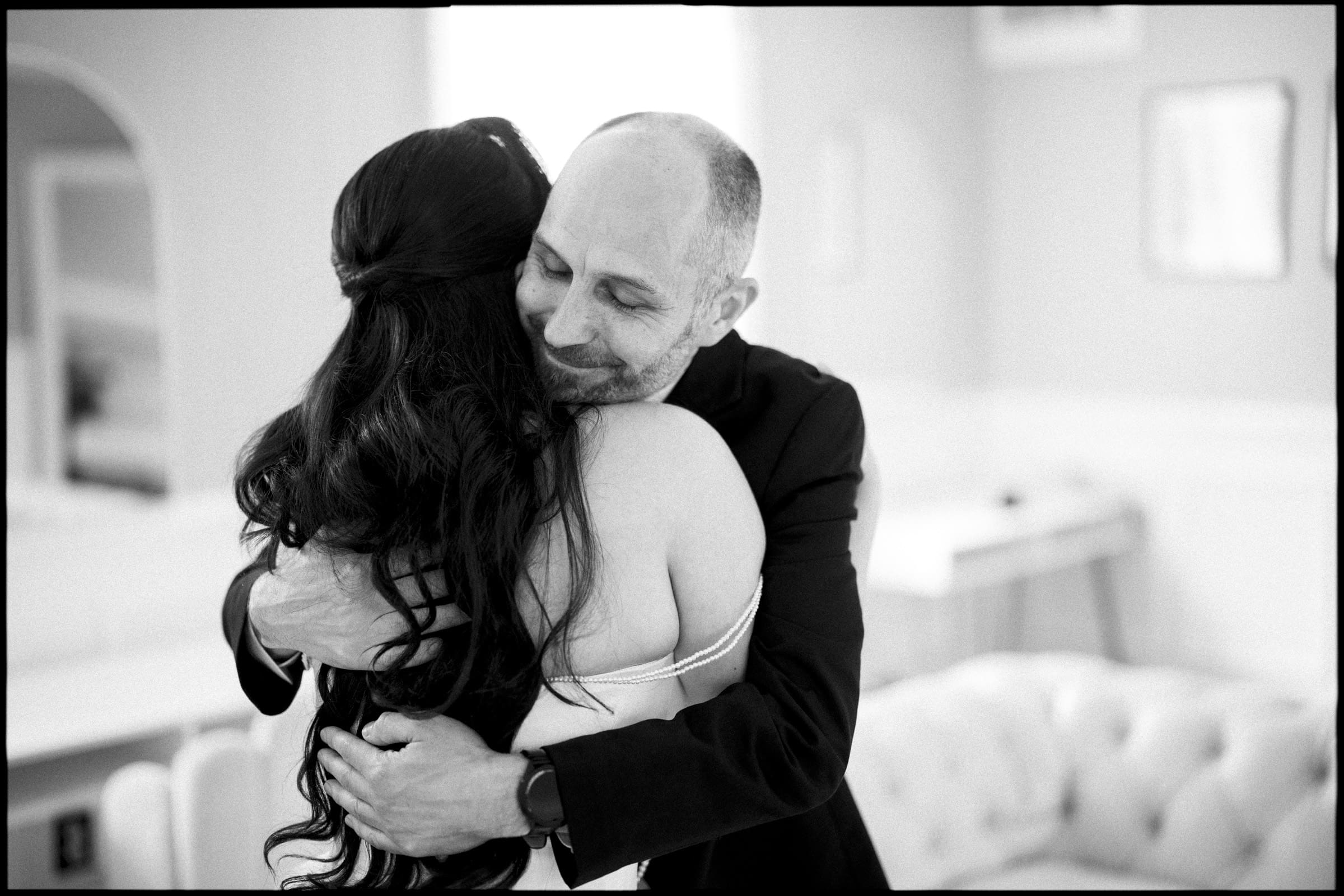 A man with a bald head and a beard warmly hugs a woman with long dark hair in an indoor setting at a Wakefield Barn wedding, both appearing joyful and emotional. The scene is in black and white. By award-winning Raleigh wedding photographer, Joe Payne.