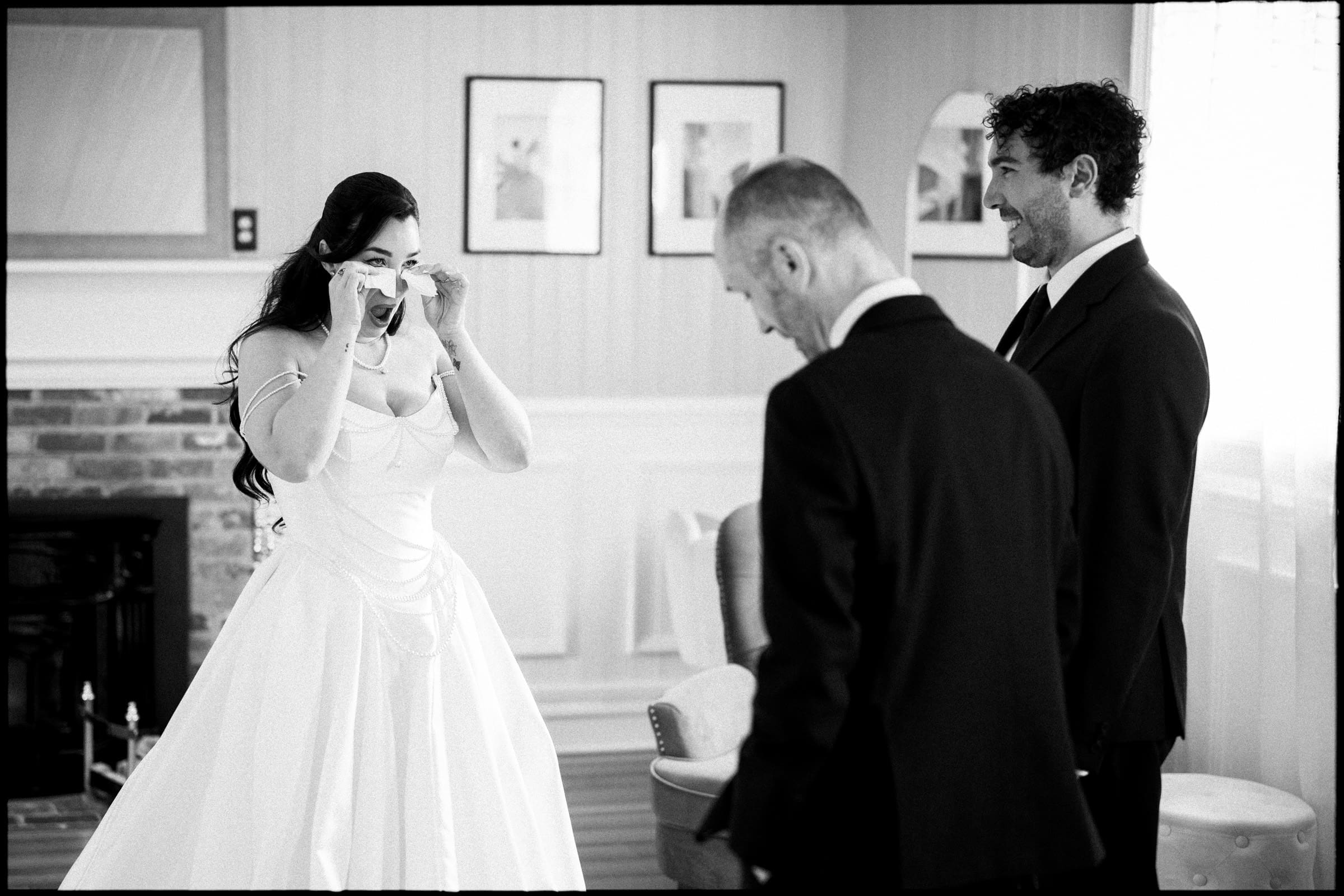 A bride in a white dress wipes her eyes as two men in suits stand nearby in a bright room, sharing an emotional moment before the historic wedding of Keiko and Ryan at Wakefield Barn. By award-winning Raleigh wedding photographer, Joe Payne.