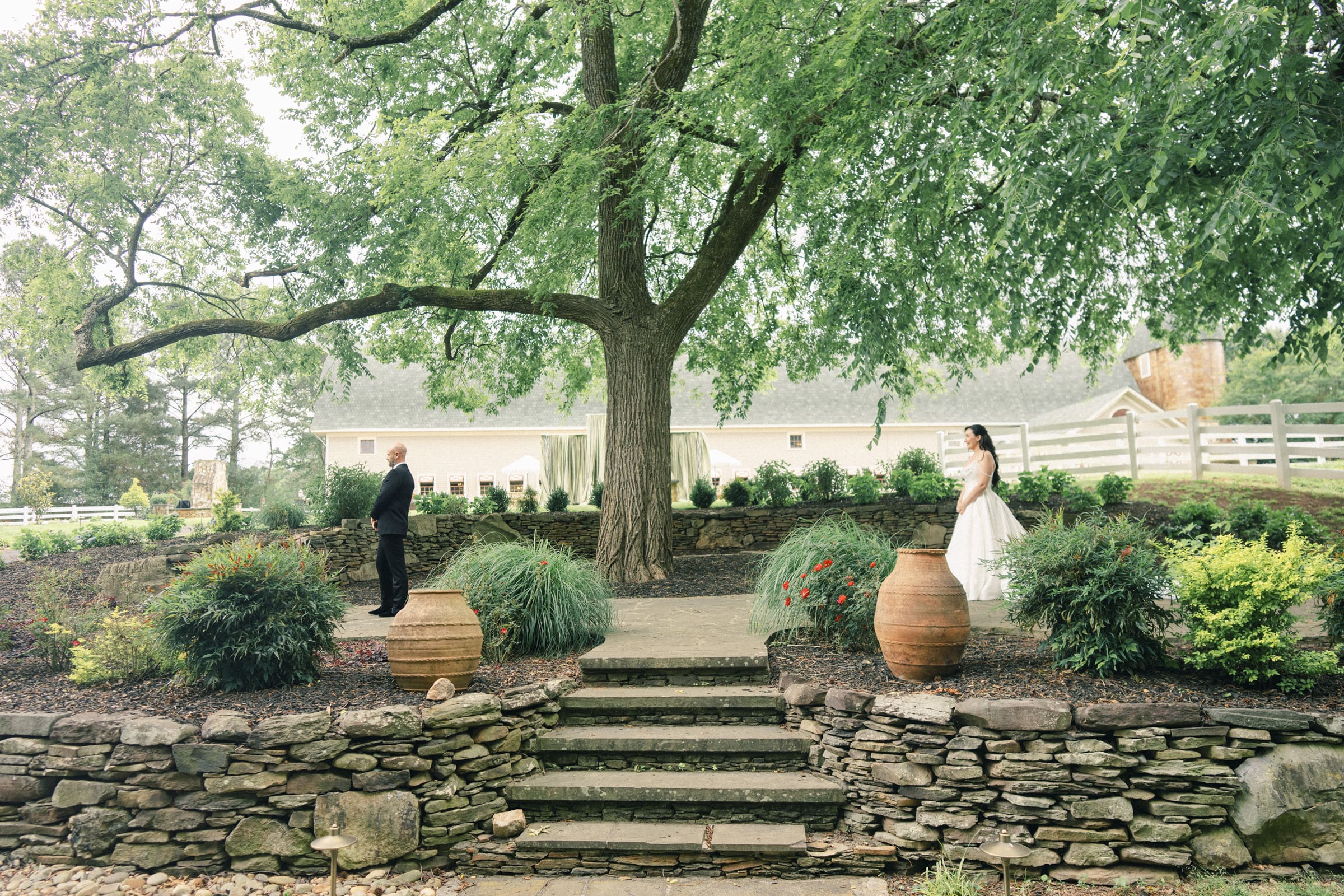 A bride and groom stand apart under a large tree in a landscaped garden at their Wakefield Barn wedding, facing each other across stone steps, surrounded by greenery with a rustic building and white fence in the background. By award-winning Raleigh wedding photographer, Joe Payne.