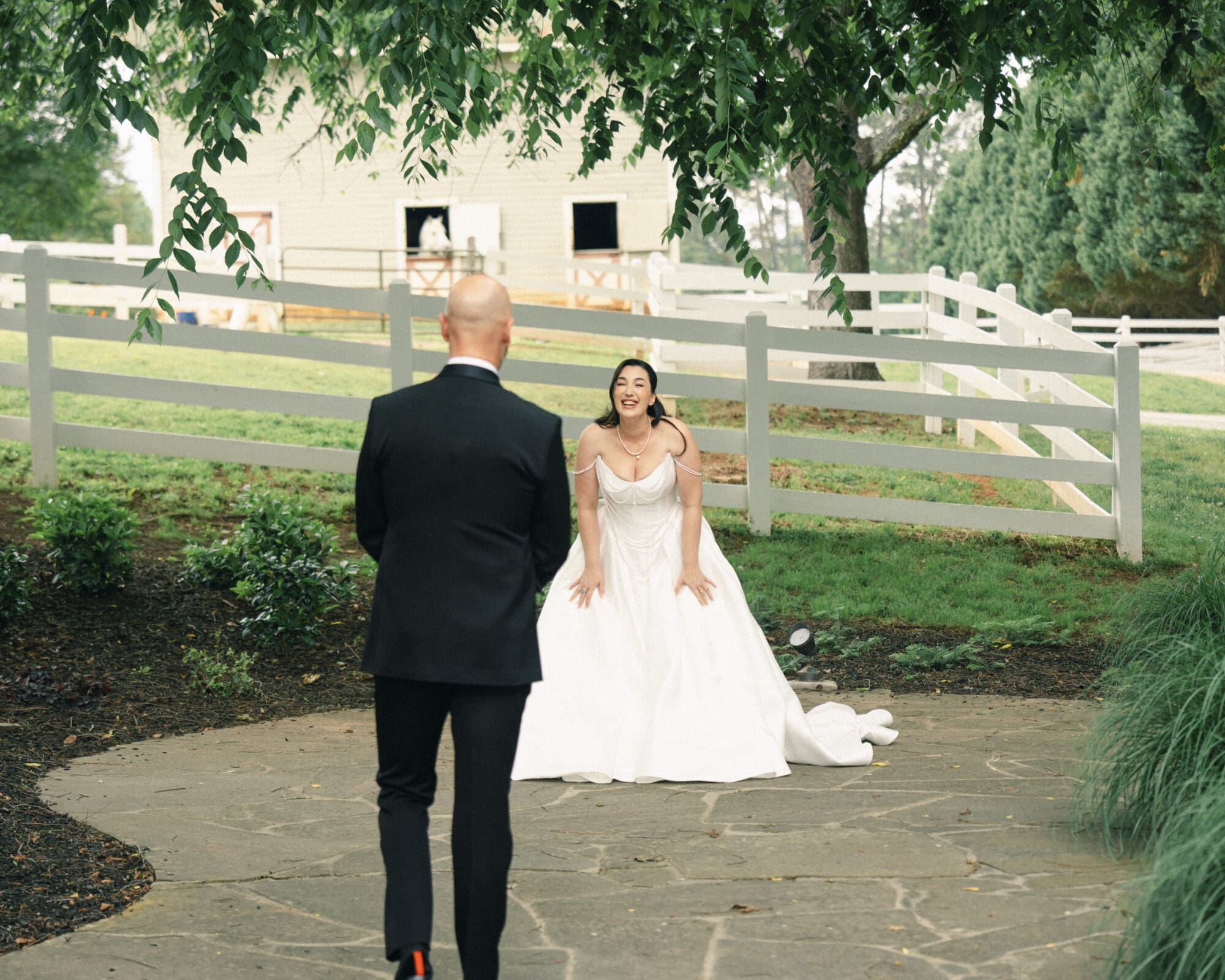 A bride in a white gown smiles joyfully as she sees a man in a black suit approaching her outdoors near a white fence and green trees at their romantic Wakefield Barn wedding. By award-winning Raleigh wedding photographer, Joe Payne.