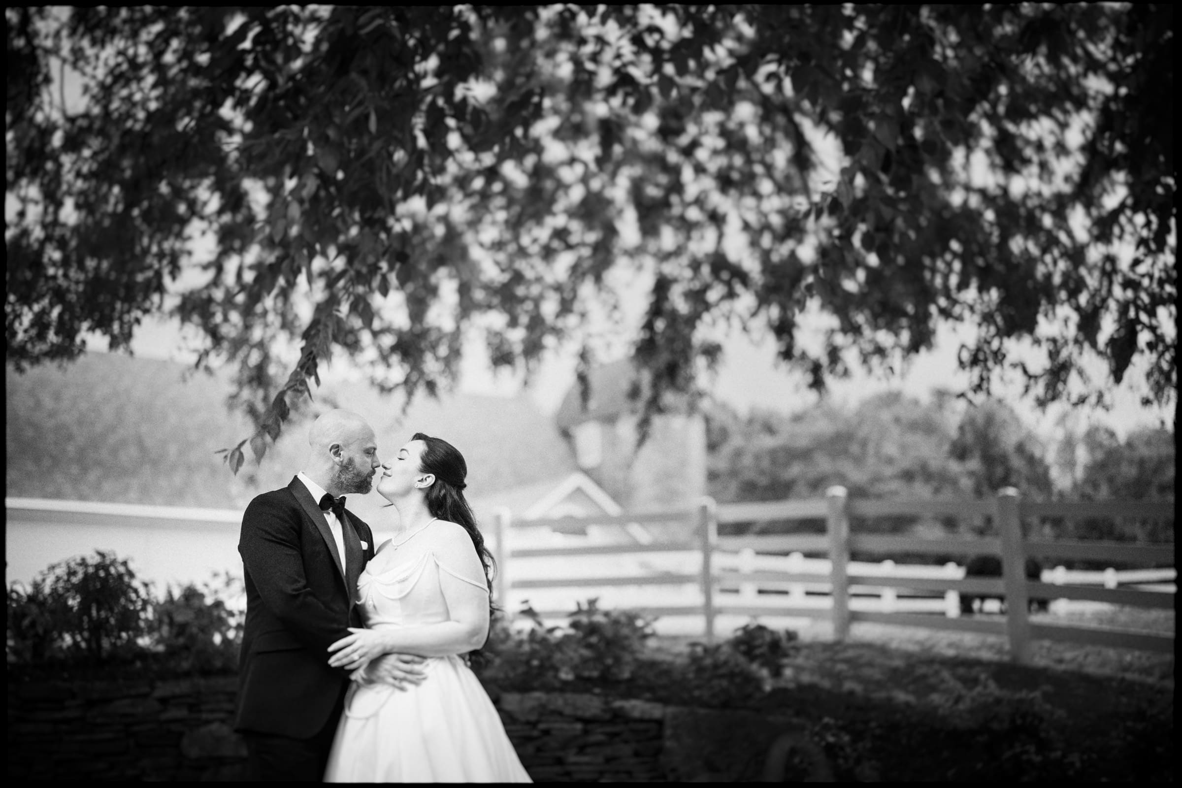 A bride and groom stand close together outdoors at their Wakefield Barn wedding, smiling and looking into each other’s eyes beneath leafy tree branches, with a fence and buildings blurred in the background. By award-winning Raleigh wedding photographer, Joe Payne.