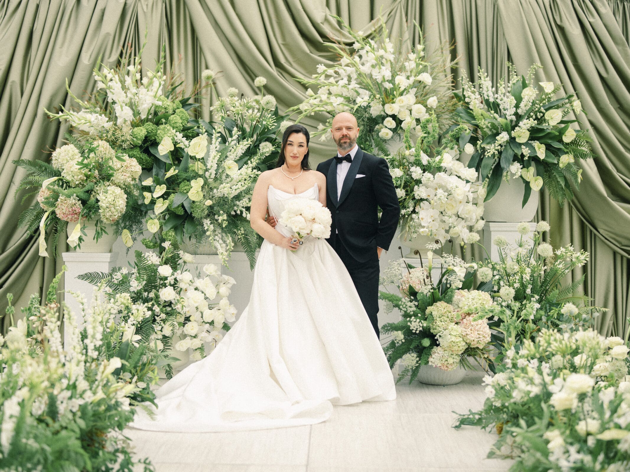 A bride in a white gown and a groom in a dark tuxedo stand together at their Historic Wakefield Barn Wedding, surrounded by lush white and green floral arrangements and draped green fabric. By award-winning Raleigh wedding photographer, Joe Payne.