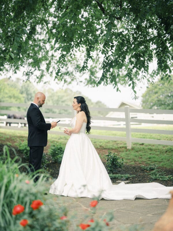 A bride in a white gown and groom in a black suit stand outdoors under leafy trees at their Wakefield Barn wedding, facing each other as the groom reads from a piece of paper. A white fence and red flowers complete this picturesque scene. By award-winning Raleigh wedding photographer, Joe Payne.