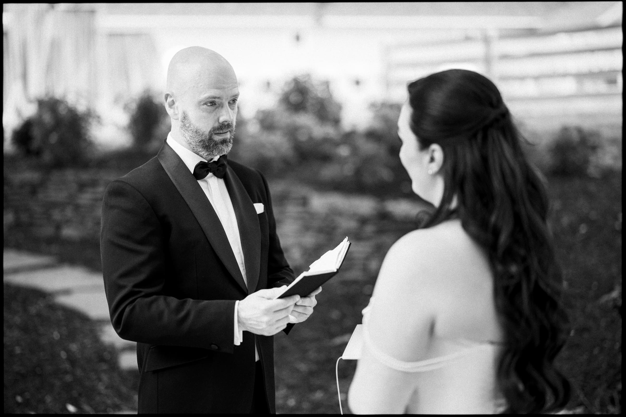 A man in a tuxedo holds a book, facing a woman in a strapless dress with long hair during an outdoor wakefield barn wedding ceremony. The black and white image captures the elegance of this historic wedding moment. By award-winning Raleigh wedding photographer, Joe Payne.