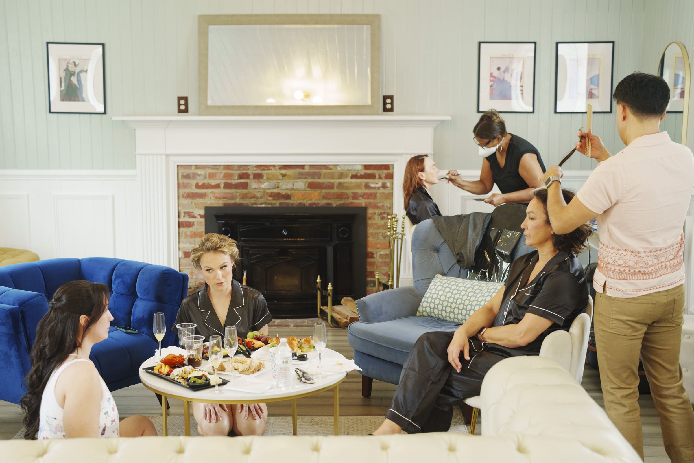 Four women in pajamas relax in a cozy living room, chatting and snacking, while two stylists do their hair and makeup. The casual, cheerful scene has a fireplace in the background—perfect pre-wedding vibes for a Wakefield barn wedding. By award-winning Raleigh wedding photographer, Joe Payne.