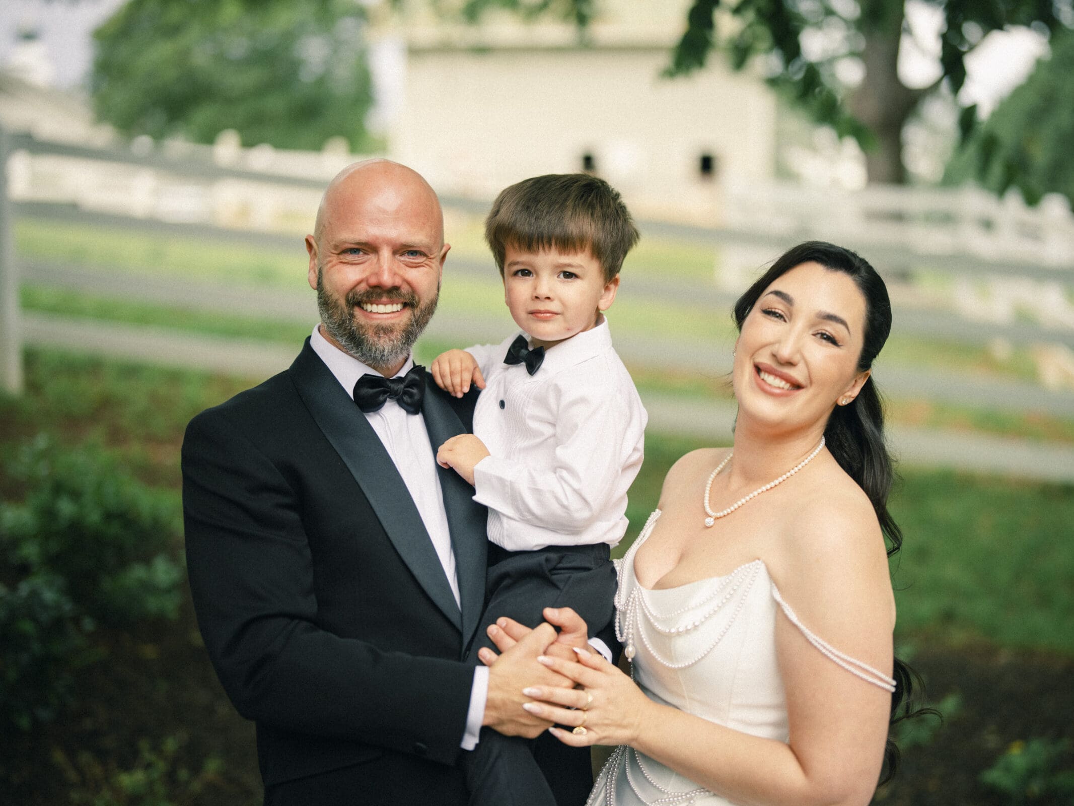 A smiling man in a black tuxedo, a woman in an elegant white dress, and a young boy in dress clothes pose together outdoors in front of a white fence and greenery at Keiko & Ryan’s historic Wakefield Barn wedding. By award-winning Raleigh wedding photographer, Joe Payne.