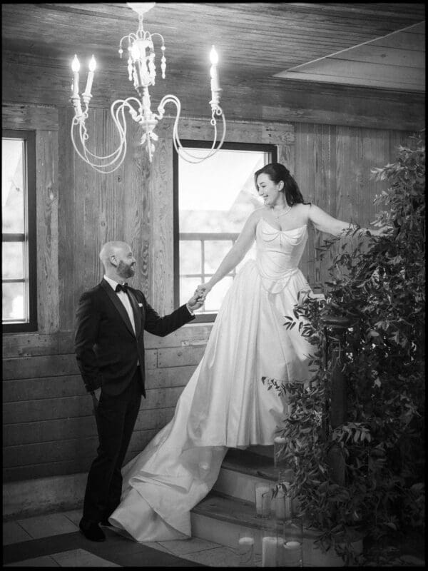 A groom in a black tuxedo holds the hand of a bride in a long, elegant white gown as she stands on stairs. They gaze at each other in a rustic room with wood walls and large windows—a perfect Wakefield Barn wedding setting. By award-winning Raleigh wedding photographer, Joe Payne.
