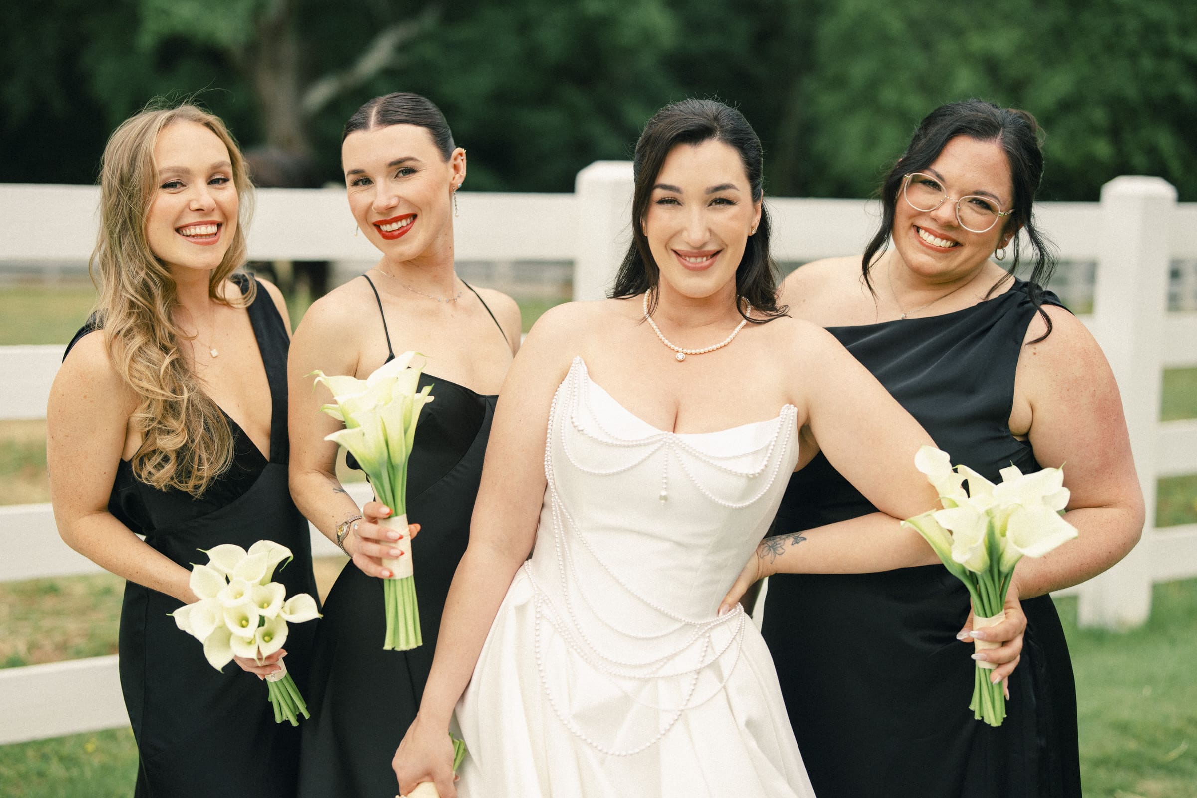 A bride in a white dress stands smiling with three bridesmaids in black dresses, all holding white calla lilies, in front of a white fence and green trees at a picturesque Wakefield Barn wedding. By award-winning Raleigh wedding photographer, Joe Payne.