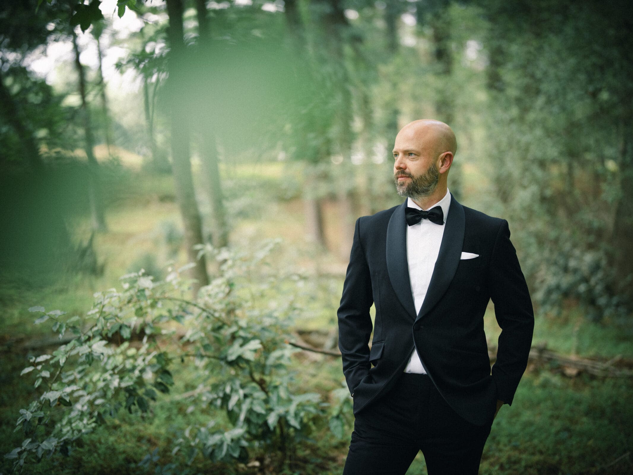 A man in a black tuxedo with a bow tie stands in a lush, green forest, looking to the side with his hands in his pockets—a serene moment from Keiko and Ryan’s Wakefield Barn wedding, with the natural scenery beautifully blurred behind him. By award-winning Raleigh wedding photographer, Joe Payne.