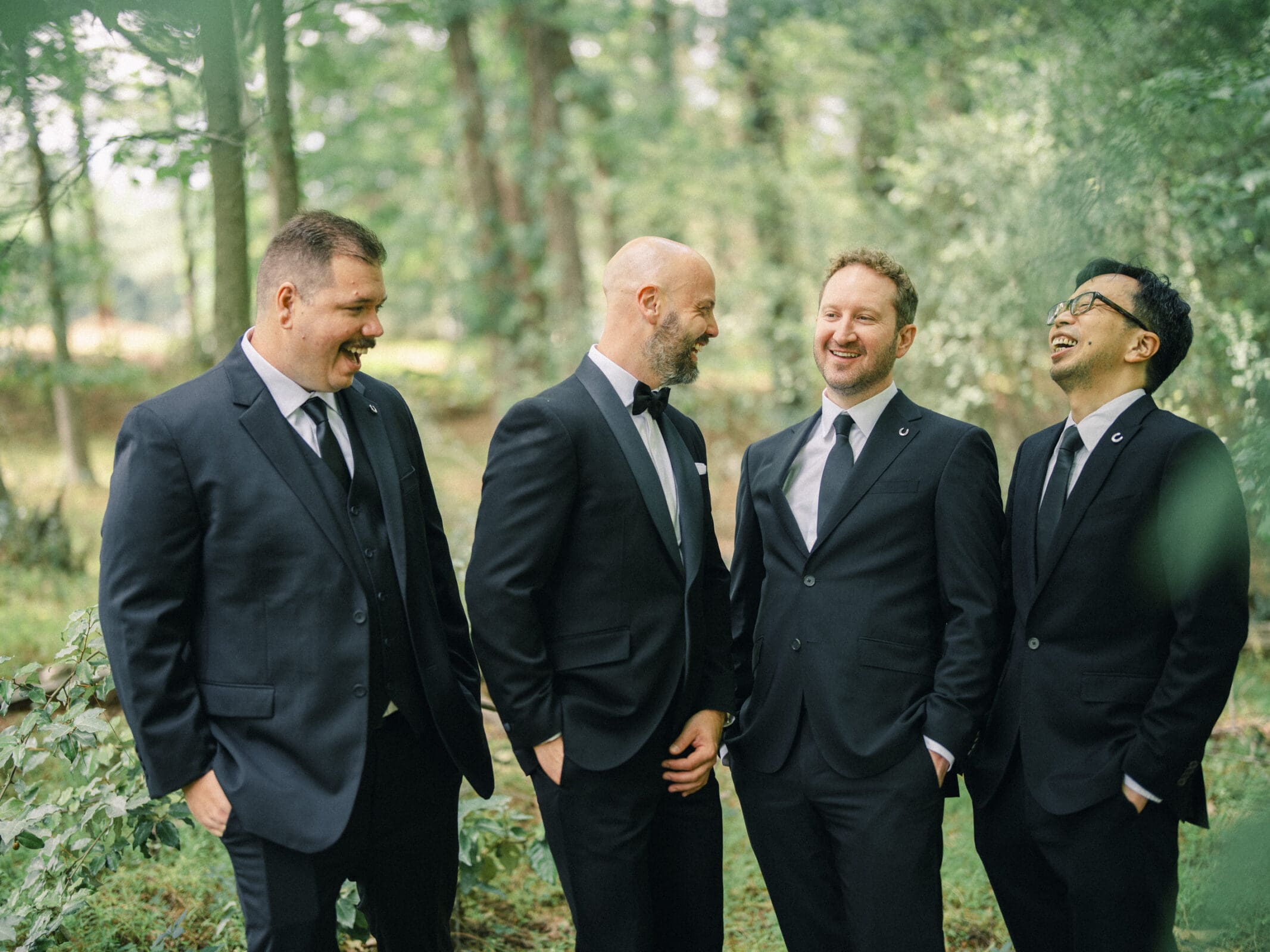 Four men in black suits stand outdoors in a green, wooded area, smiling and laughing together. They seem to be enjoying a lighthearted moment at a barn wedding, possibly at a historic wedding venue. By award-winning Raleigh wedding photographer, Joe Payne.