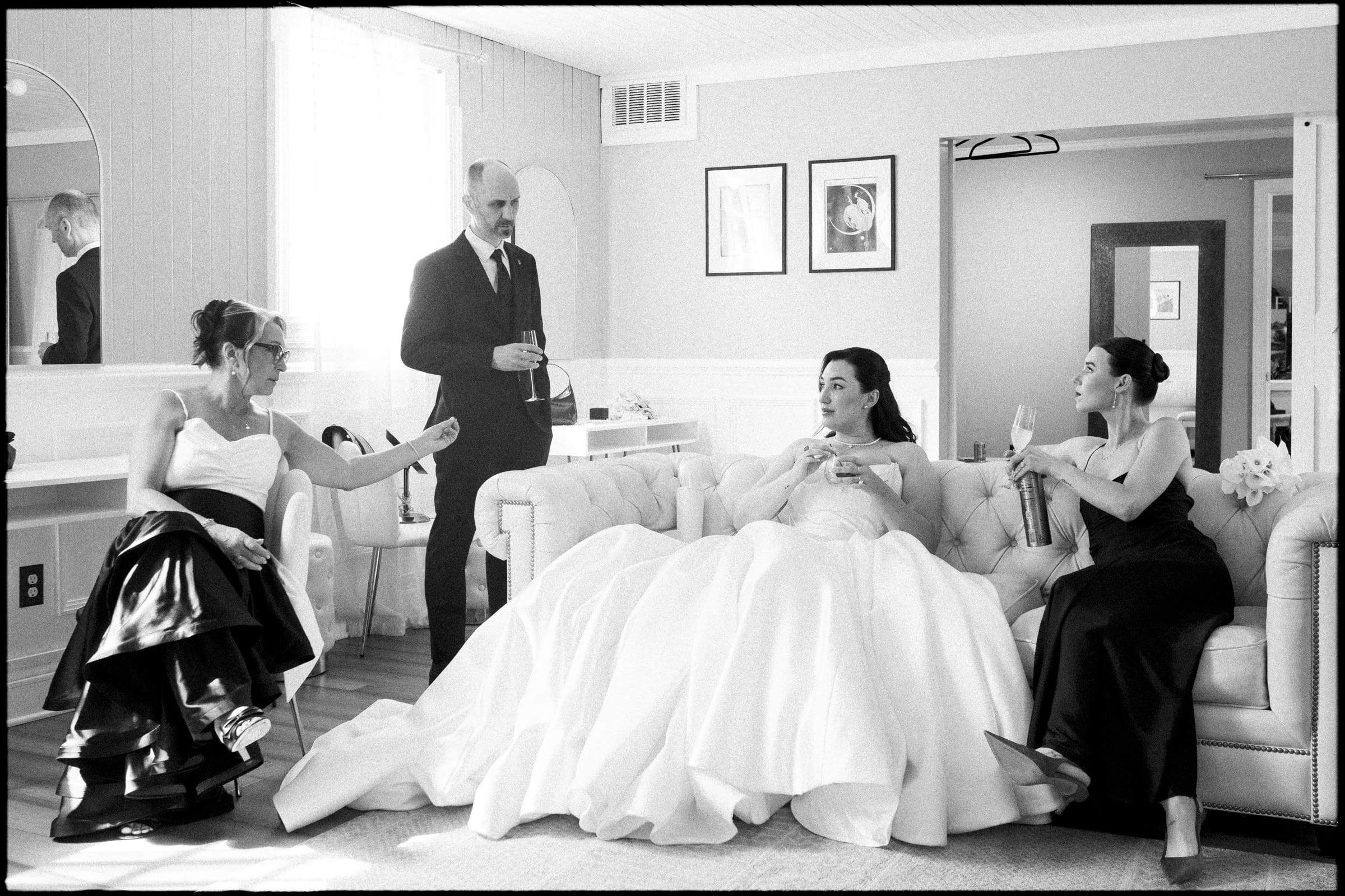 Four people in formal attire relax in a bright room at a Historic Wakefield Barn Wedding. A woman in a wedding dress sits on a sofa, chatting with two women in dark gowns and a man in a suit holding a drink. The atmosphere is calm and intimate. By award-winning Raleigh wedding photographer, Joe Payne.