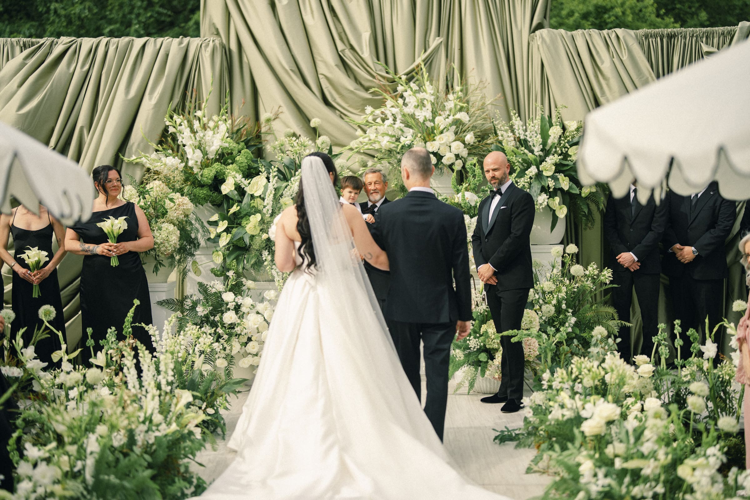 A bride in a white gown and veil walks down the aisle with a man toward the groom at an outdoor Wakefield Barn wedding, surrounded by lush greenery, white flowers, and formally dressed guests. By award-winning Raleigh wedding photographer, Joe Payne.
