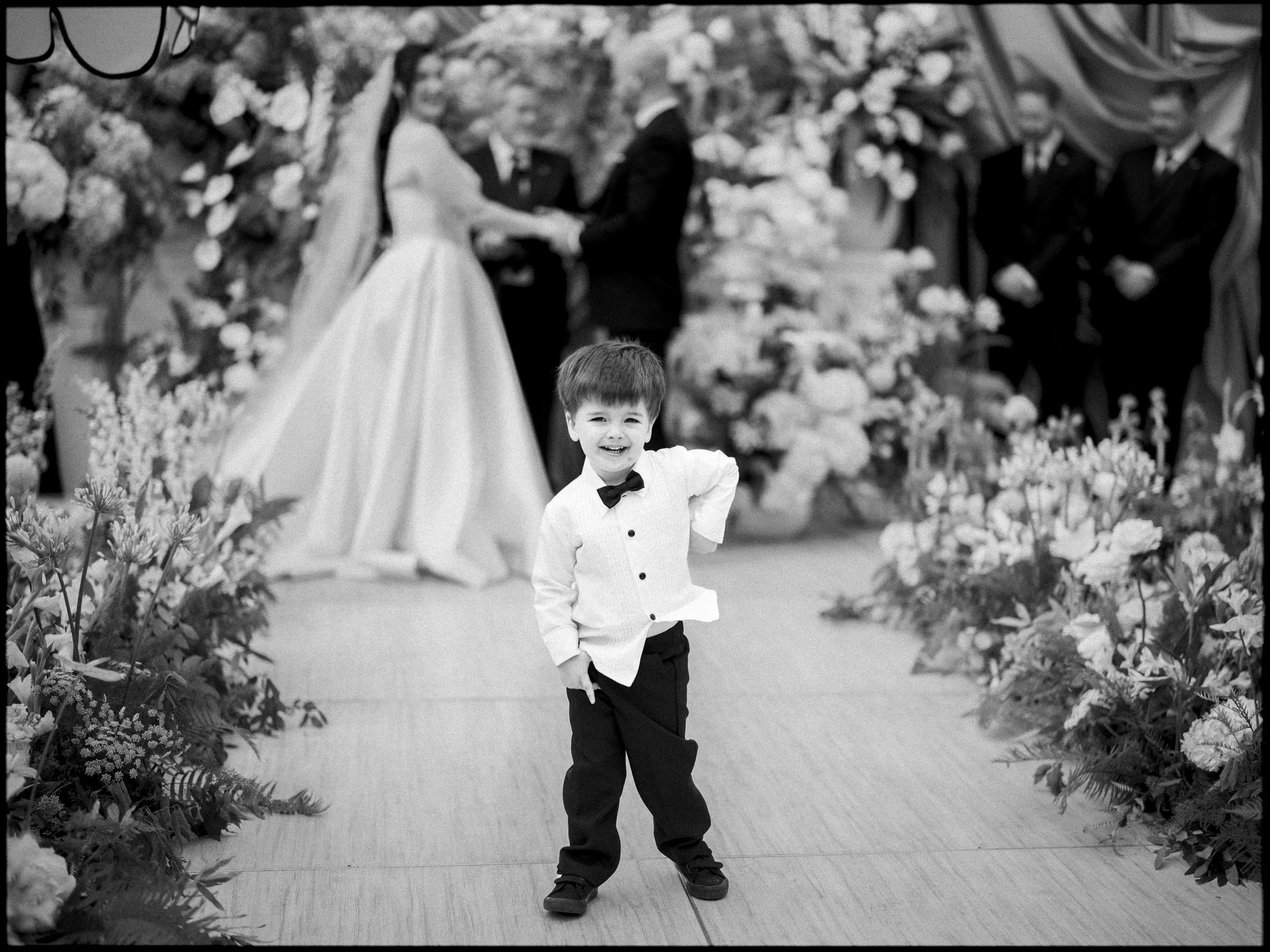 A young boy in a tuxedo smiles and poses on a floral-lined aisle at a Historic Wakefield Barn Wedding, with the bride, groom, and groomsmen blurred in the background. By award-winning Raleigh wedding photographer, Joe Payne.