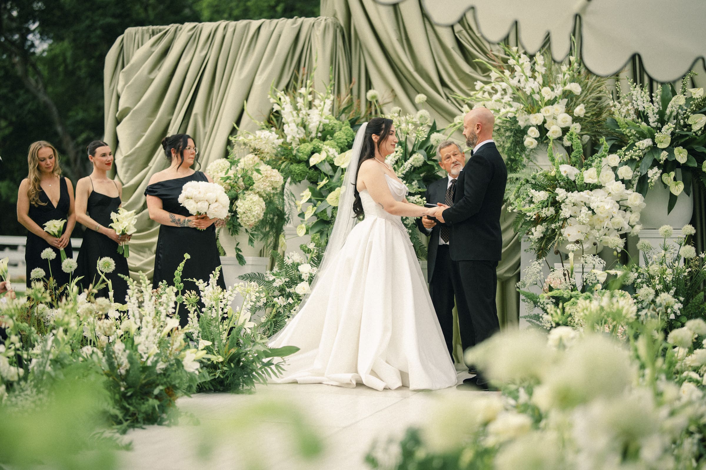 A bride and groom hold hands during their Wakefield Barn wedding ceremony, surrounded by lush white flowers and greenery. Bridesmaids in black dresses stand to the side as the officiant presides over this beautiful barn wedding scene. By award-winning Raleigh wedding photographer, Joe Payne.