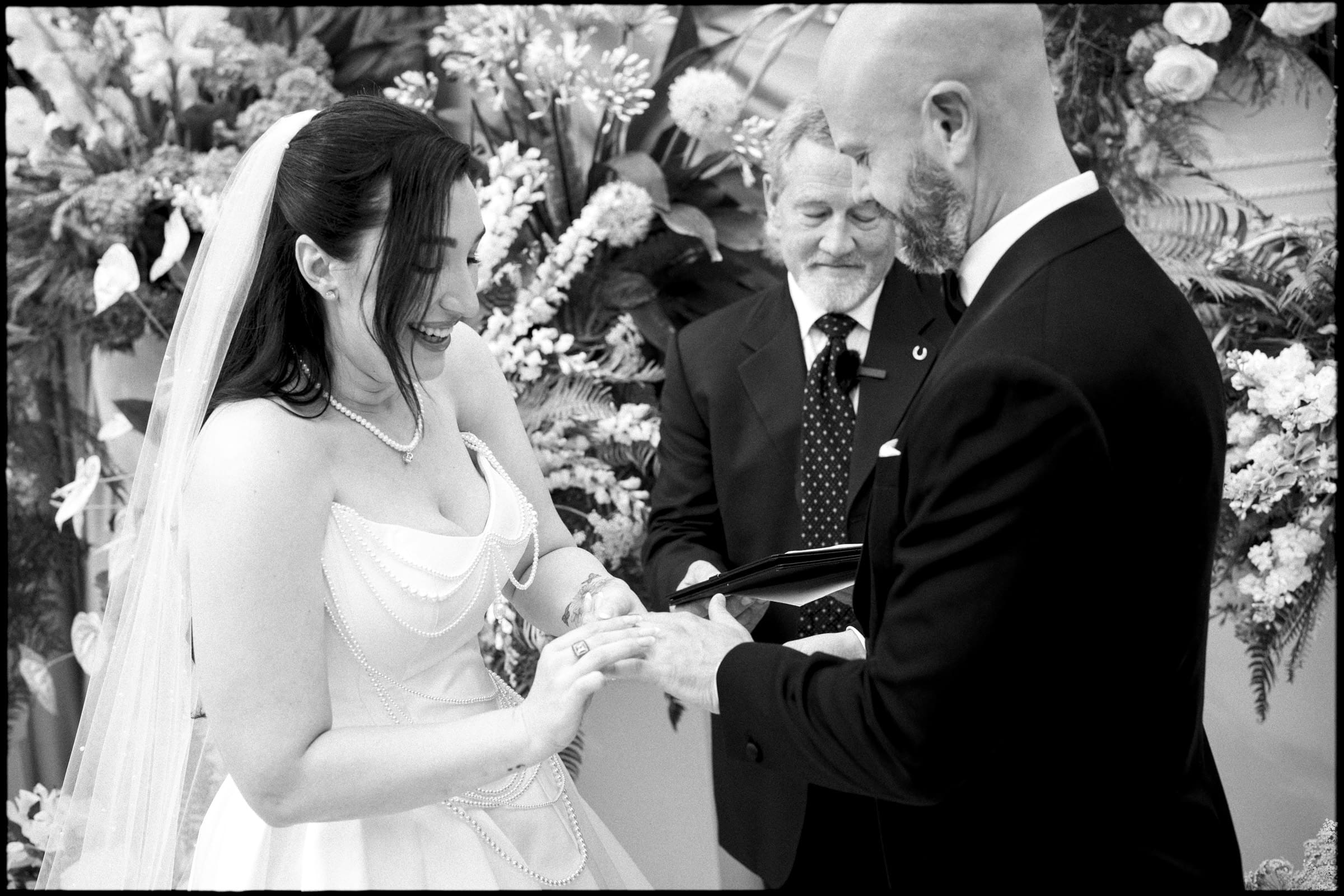A bride and groom exchange rings and smile during their Wakefield Barn wedding ceremony, with an officiant behind them, surrounded by elaborate floral arrangements. By award-winning Raleigh wedding photographer, Joe Payne.