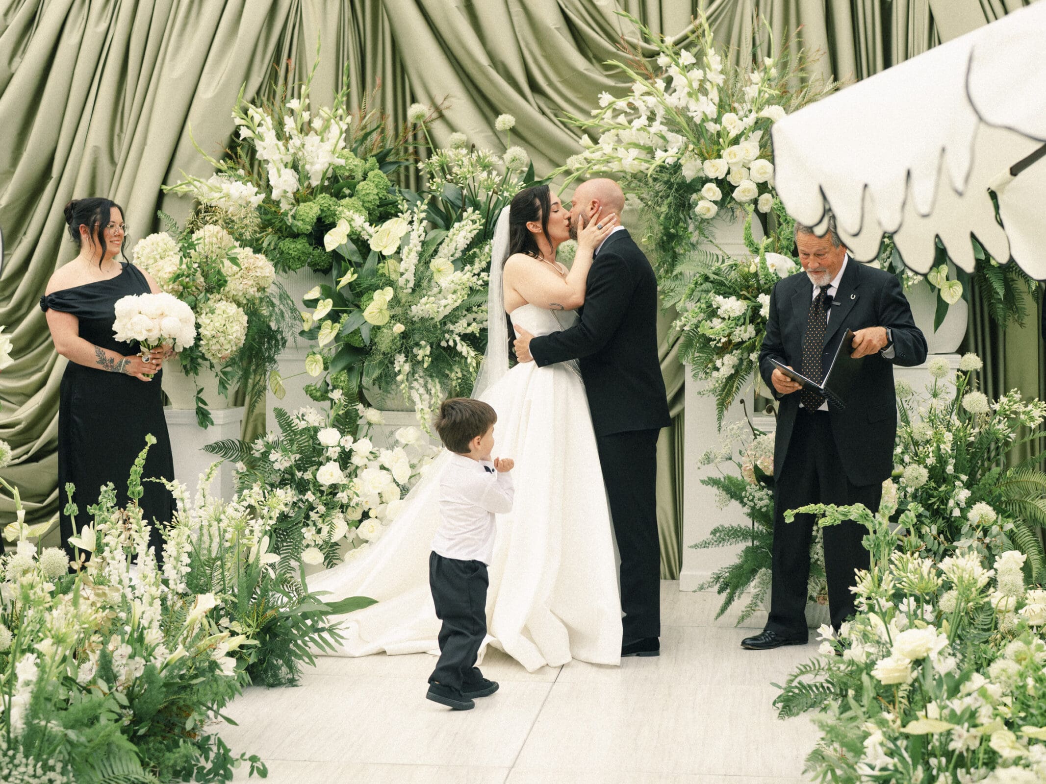 A bride and groom kiss at their Historic Wakefield Barn Wedding, surrounded by lush green and white floral arrangements, while a young boy stands in front and two adults, one holding flowers, stand nearby. By award-winning Raleigh wedding photographer, Joe Payne.