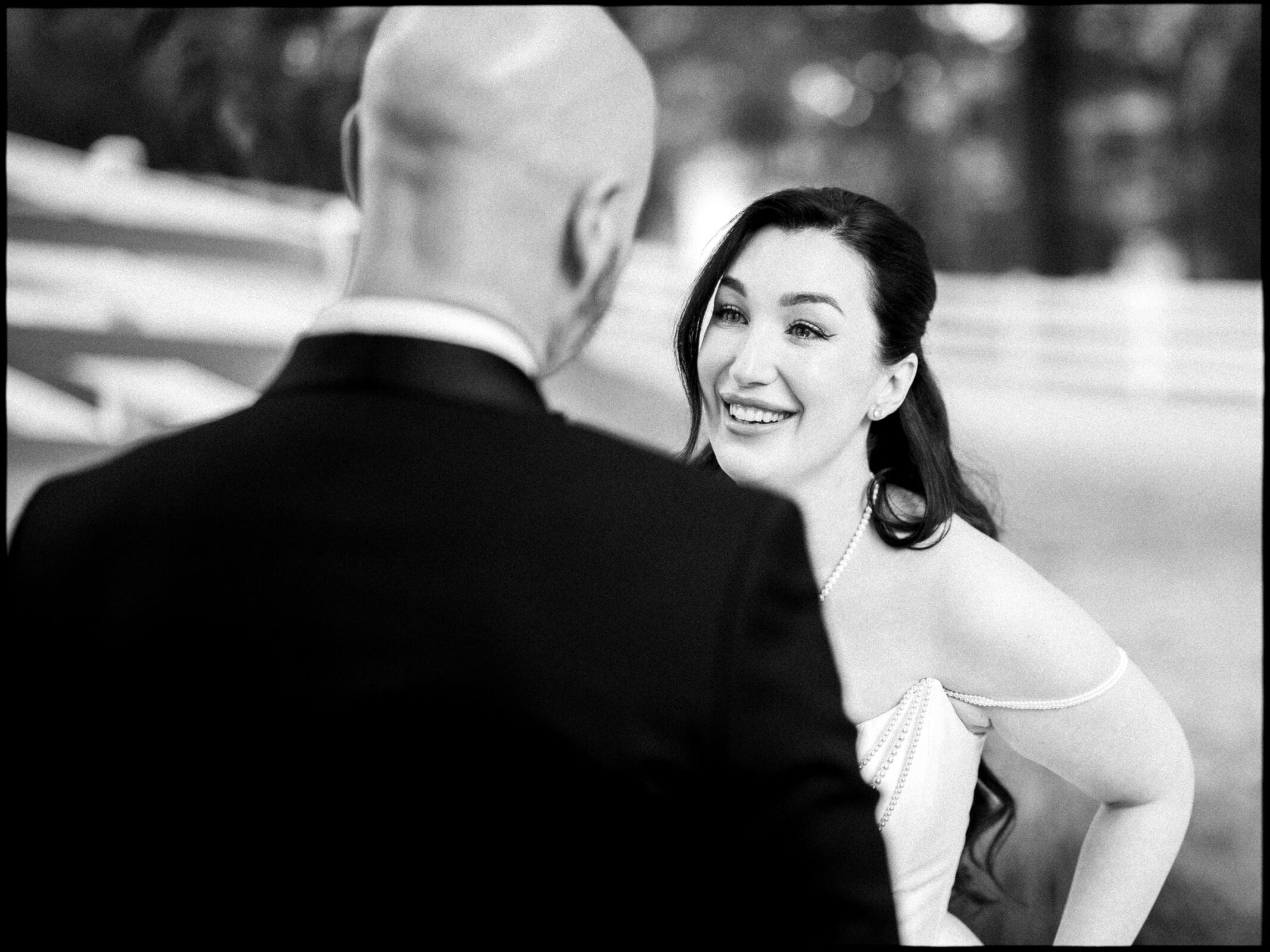 A woman in a wedding dress smiles warmly at a bald man in a suit during an outdoor moment at their Historic Wakefield Barn Wedding, with blurred trees and a white fence in the background. By award-winning Raleigh wedding photographer, Joe Payne.