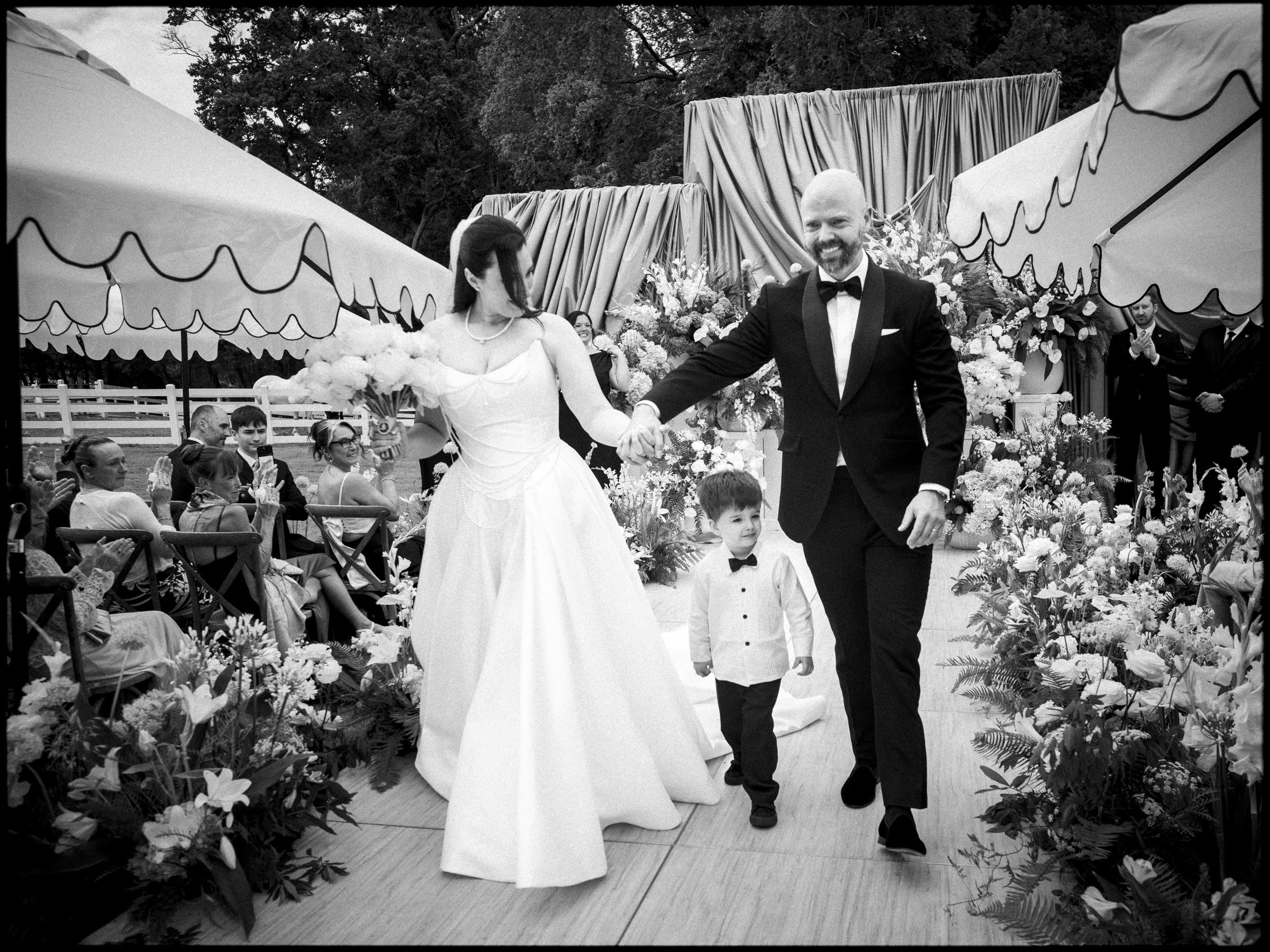 A bride and groom, holding hands with a young boy, walk down an outdoor aisle lined with flowers and guests at a Historic Wakefield Barn Wedding. The festive, black-and-white scene features decorative tents and elegantly dressed attendees. By award-winning Raleigh wedding photographer, Joe Payne.