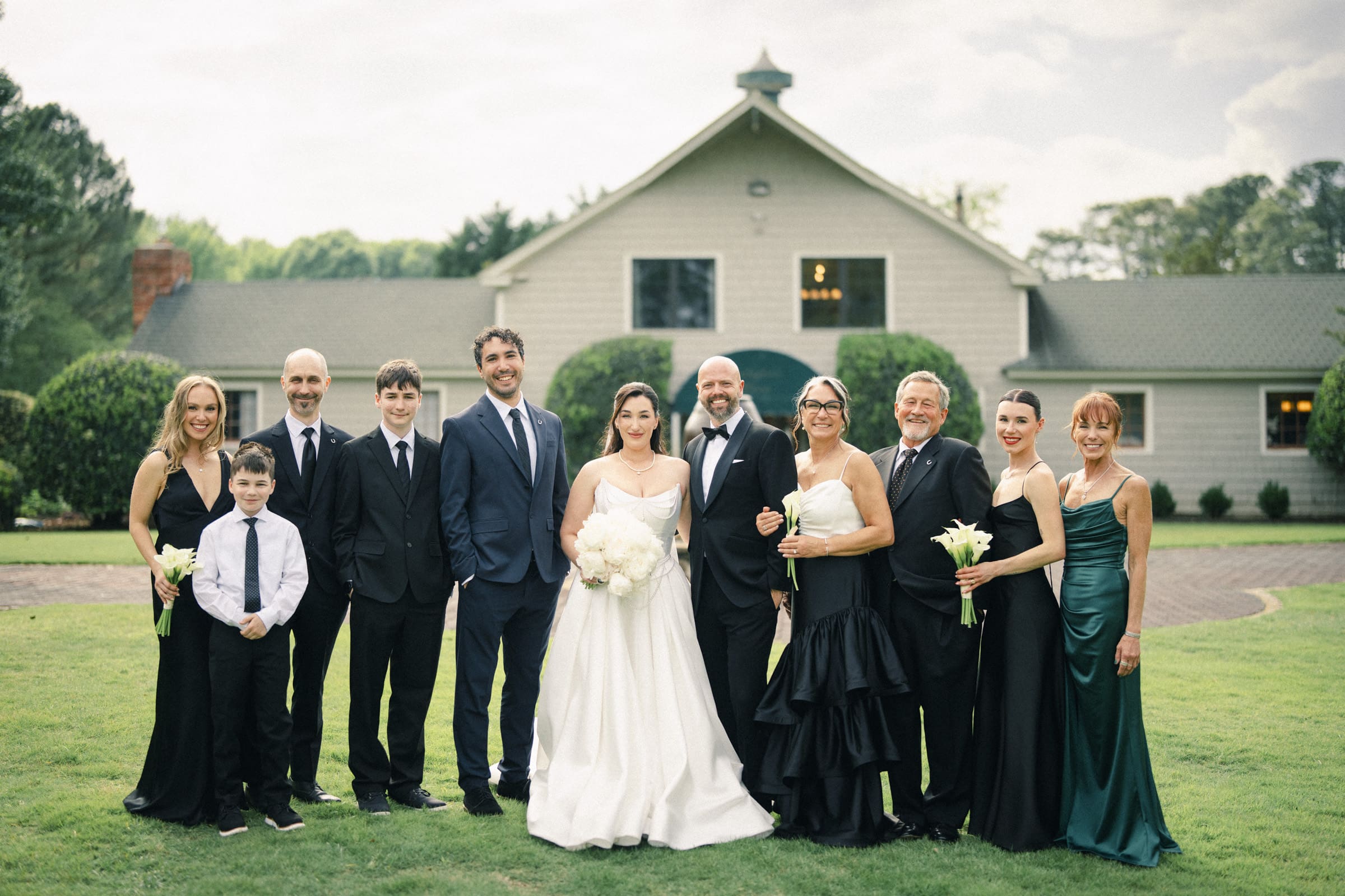 A wedding party poses outside on a lawn at a Wakefield Barn wedding. The bride in white and the groom in a dark suit stand at the center, surrounded by family and friends in formal attire, celebrating their special day at this historic venue. By award-winning Raleigh wedding photographer, Joe Payne.