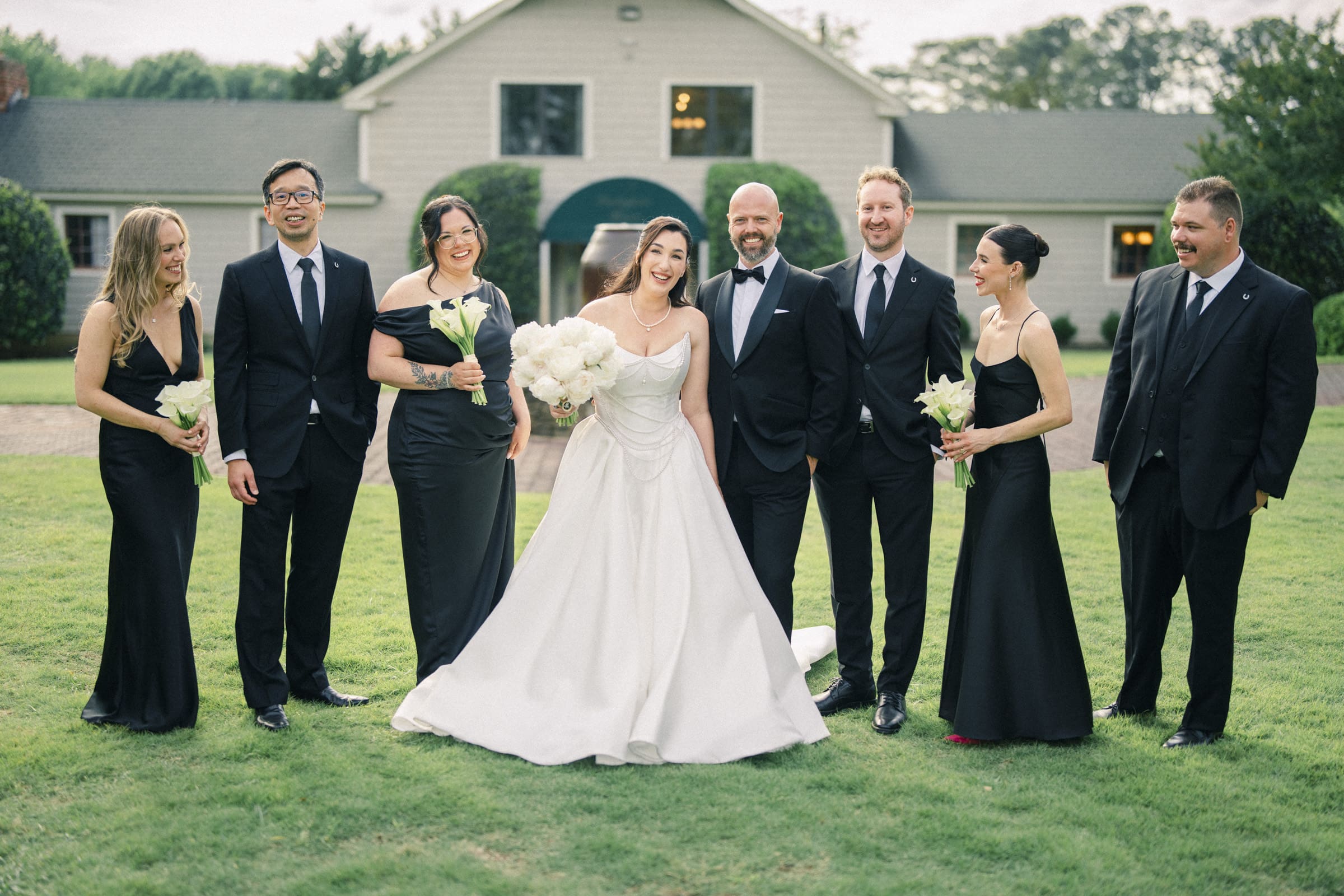 A bride in a white gown stands smiling with her bridal party, dressed in black formal attire, on a grassy lawn in front of the barn. Everyone is posing happily together, holding bouquets at this charming Wakefield Barn wedding. By award-winning Raleigh wedding photographer, Joe Payne.