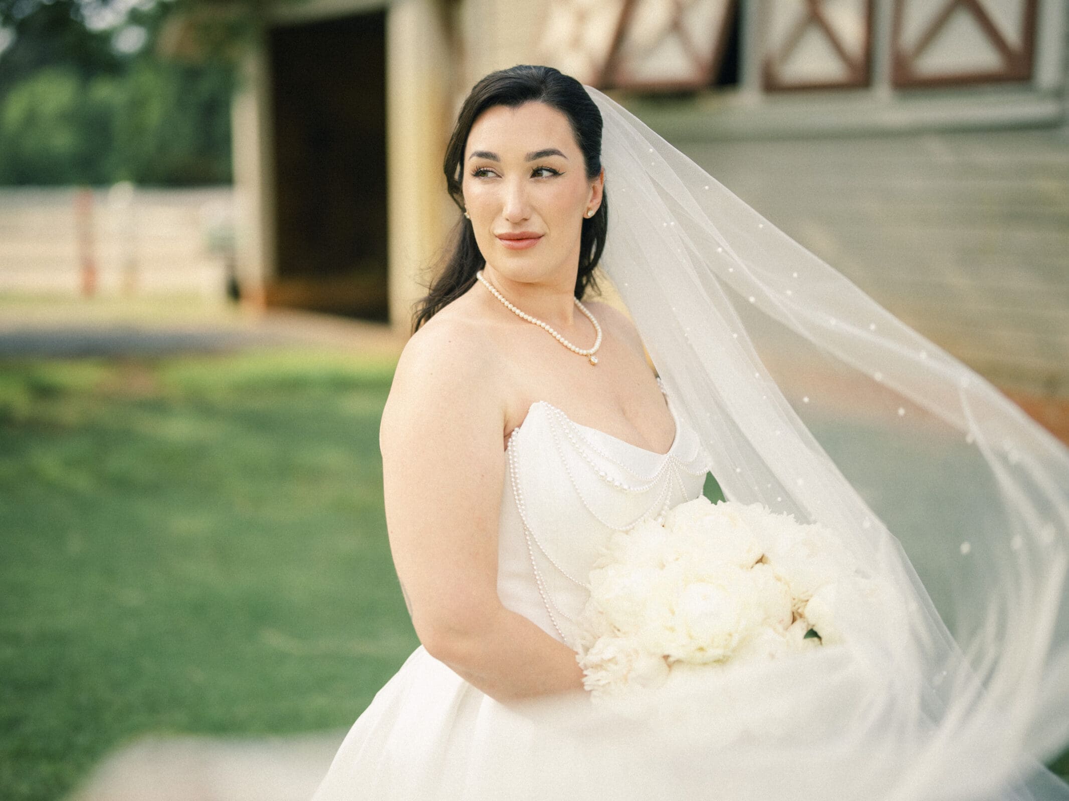 A bride in a white gown and veil stands outside, holding a bouquet of white flowers. She wears a pearl necklace and gazes to the side, with the charming greenery and rustic setting of a Wakefield barn wedding in the background. By award-winning Raleigh wedding photographer, Joe Payne.