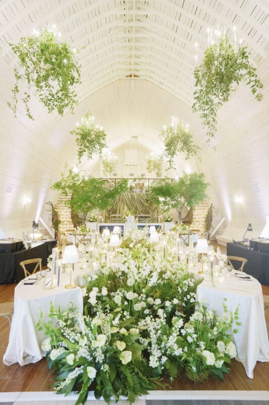 A bright, elegant Wakefield Barn wedding reception hall with white walls, hanging greenery, chandeliers, and lush floral arrangements of white flowers and greenery on and around the dining tables. By award-winning Raleigh wedding photographer, Joe Payne.