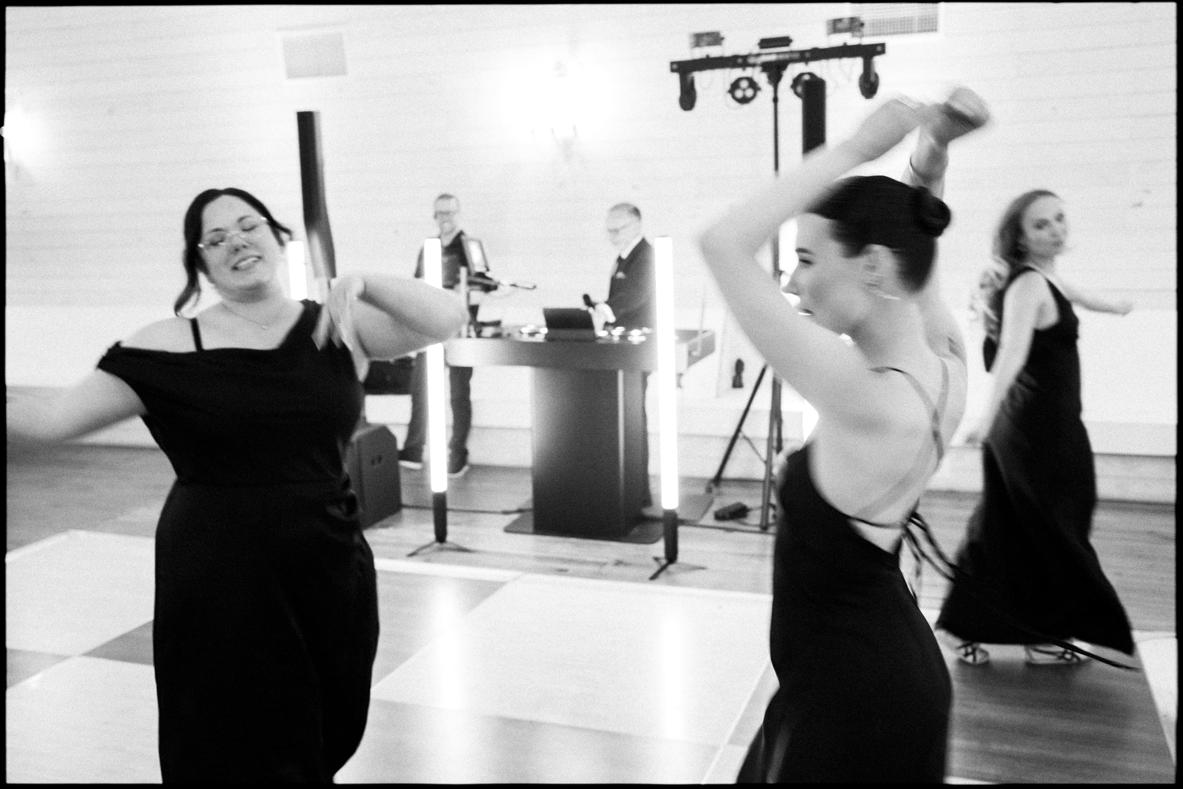 Three women in black dresses dance energetically on a ballroom floor, while two DJs stand behind the booth. The lively black-and-white scene captures the joy of a wakefield barn wedding, making it a night to remember. By award-winning Raleigh wedding photographer, Joe Payne.