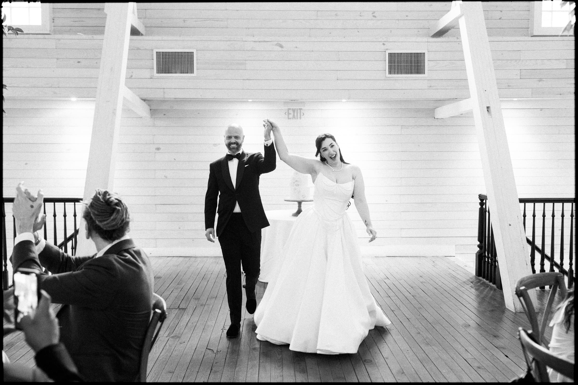 A newlywed couple, dressed in a tuxedo and white gown, joyfully enter a bright, airy Wakefield barn wedding venue holding hands as guests applaud. The moment feels celebratory and filled with happiness. By award-winning Raleigh wedding photographer, Joe Payne.