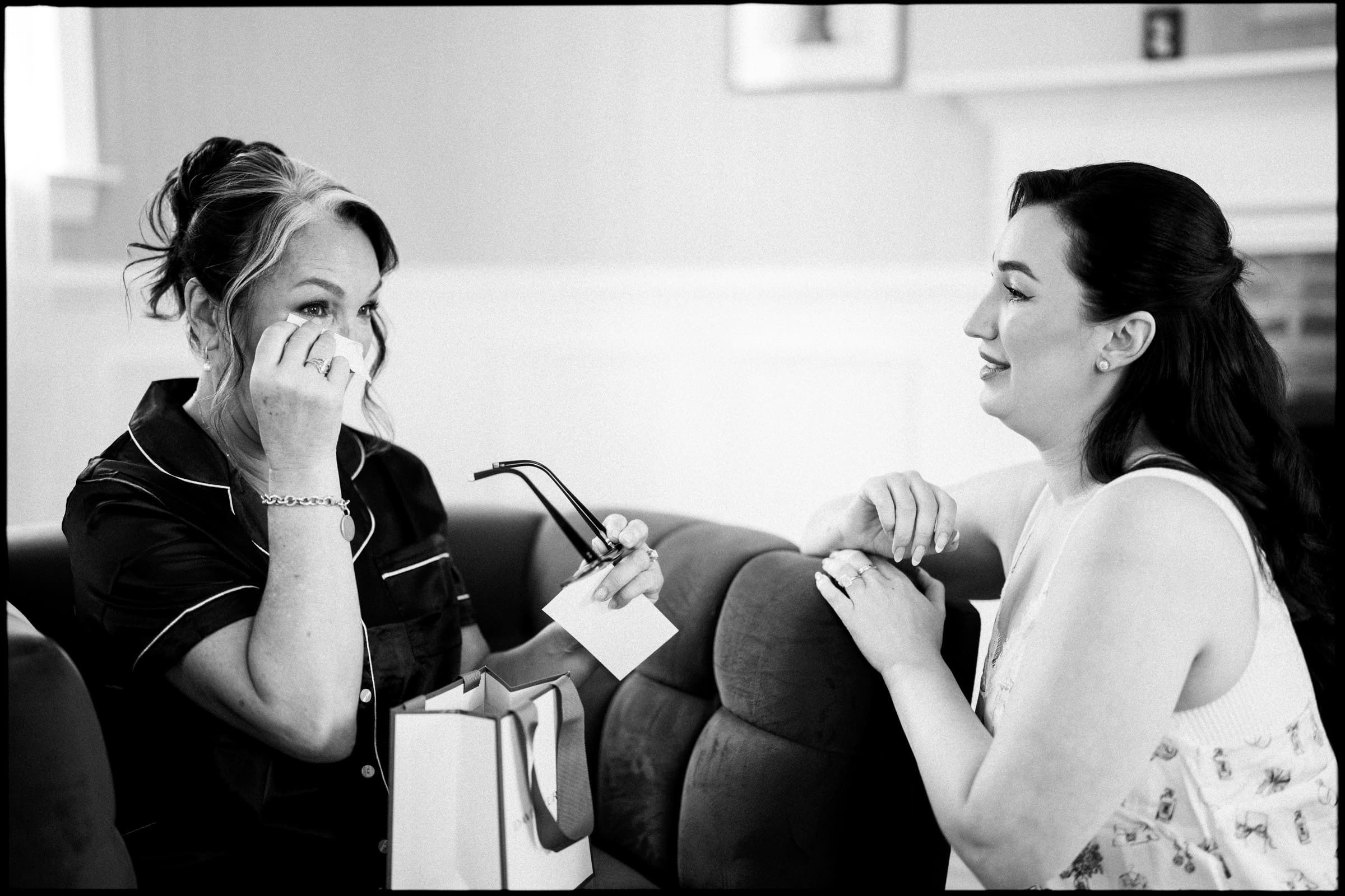A woman wipes tears from her eyes while holding glasses and a tissue. Another woman sits beside her, smiling gently, as they share an emotional moment on a sofa during Keiko and Ryan’s historic barn wedding. By award-winning Raleigh wedding photographer, Joe Payne.
