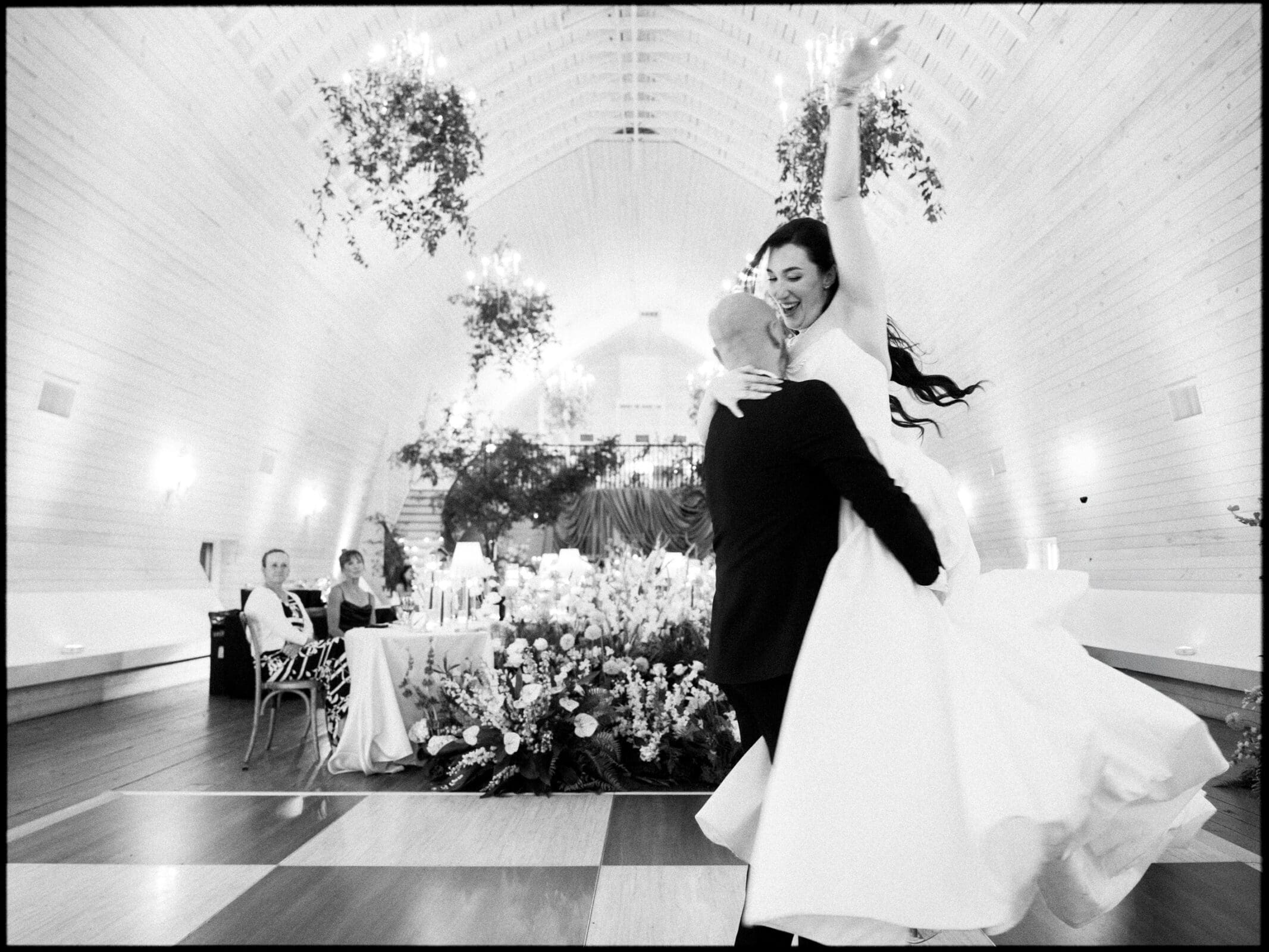 A groom lifts his smiling bride as they dance in an elegant, flower-filled Wakefield Barn wedding venue with high arched ceilings, while guests watch from tables in the background. By award-winning Raleigh wedding photographer, Joe Payne.