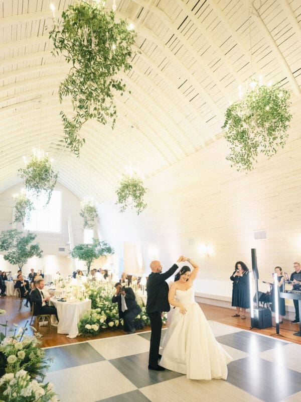A bride and groom share their first dance in a bright, elegant hall with high white ceilings, hanging greenery, and guests at decorated tables. Musicians perform nearby as natural light fills this beautiful historic wedding venue. By award-winning Raleigh wedding photographer, Joe Payne.