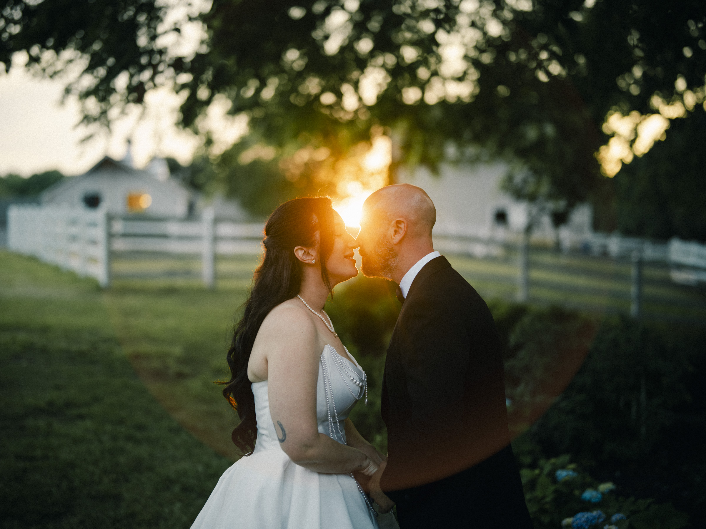 A bride and groom stand close together outdoors at sunset, touching noses and smiling, with a fence, trees, and the Wakefield Barn in the background as soft sunlight creates a romantic glow at Keiko and Ryan's Historic Wakefield Barn wedding. By award-winning Raleigh wedding photographer, Joe Payne.