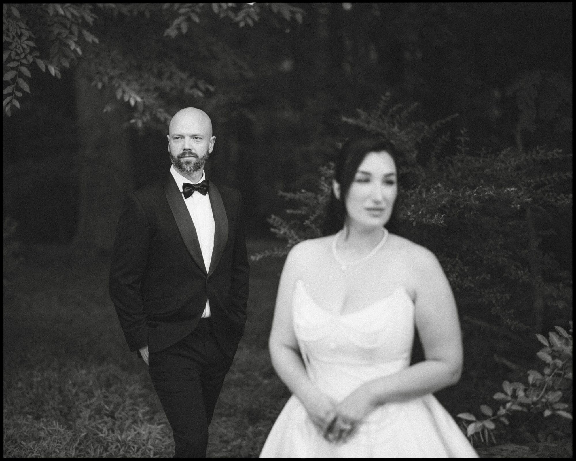 A black and white photo of a man in a tuxedo stands in the background, looking ahead, as a woman in a wedding dress stands out of focus amid trees—capturing the timeless romance of a Wakefield Barn wedding. By award-winning Raleigh wedding photographer, Joe Payne.