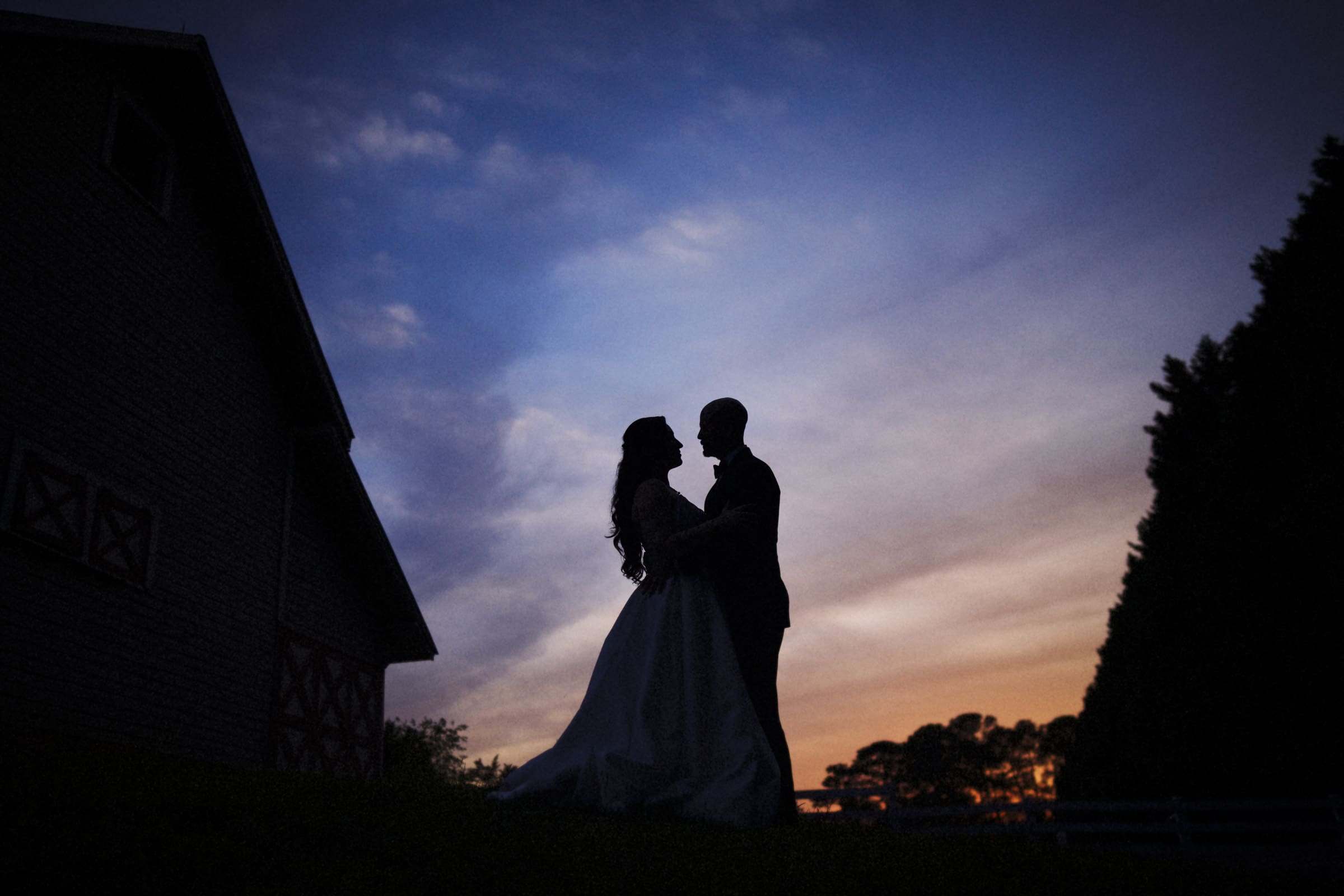 Silhouette of a bride and groom embracing outdoors at sunset, with a barn and trees in the background and a colorful sky—perfect for a romantic Wakefield Barn wedding. By award-winning Raleigh wedding photographer, Joe Payne.