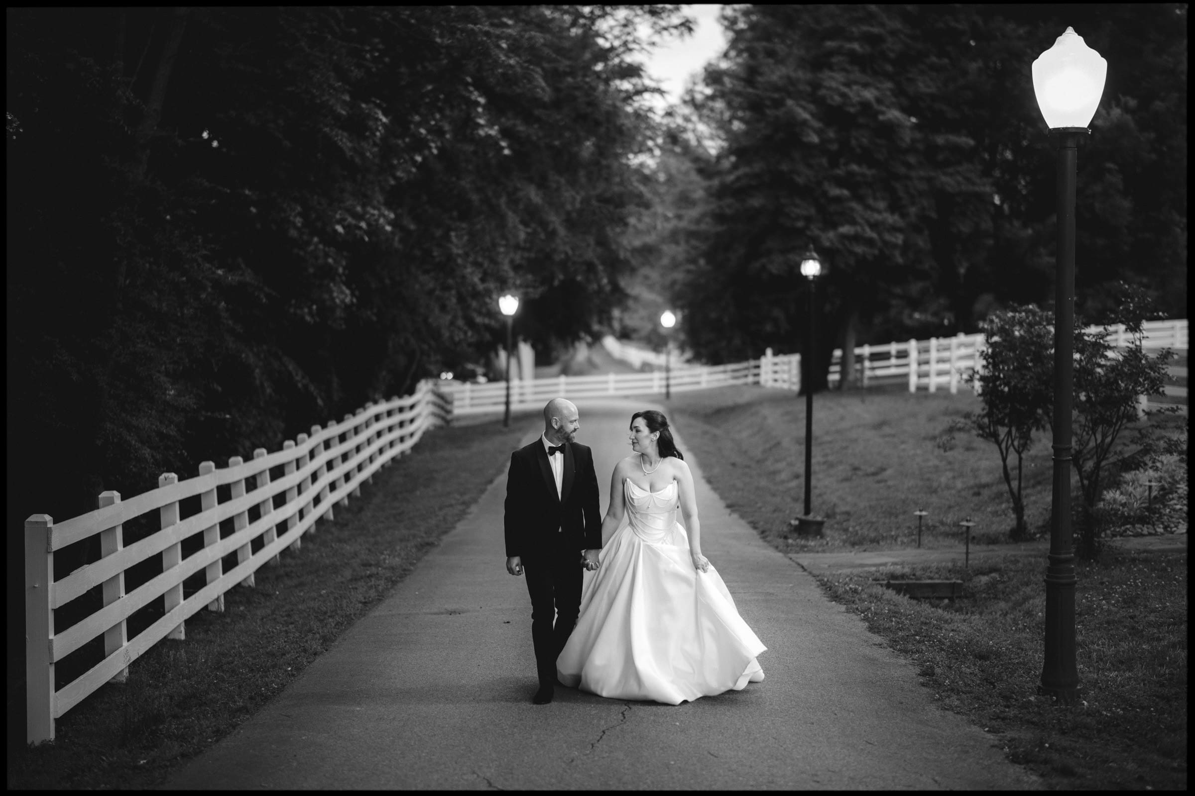 A bride and groom walk hand in hand down a tree-lined path with white fences and street lamps, smiling at each other. The black-and-white scene creates a classic, romantic atmosphere at this Wakefield Barn wedding. By award-winning Raleigh wedding photographer, Joe Payne.