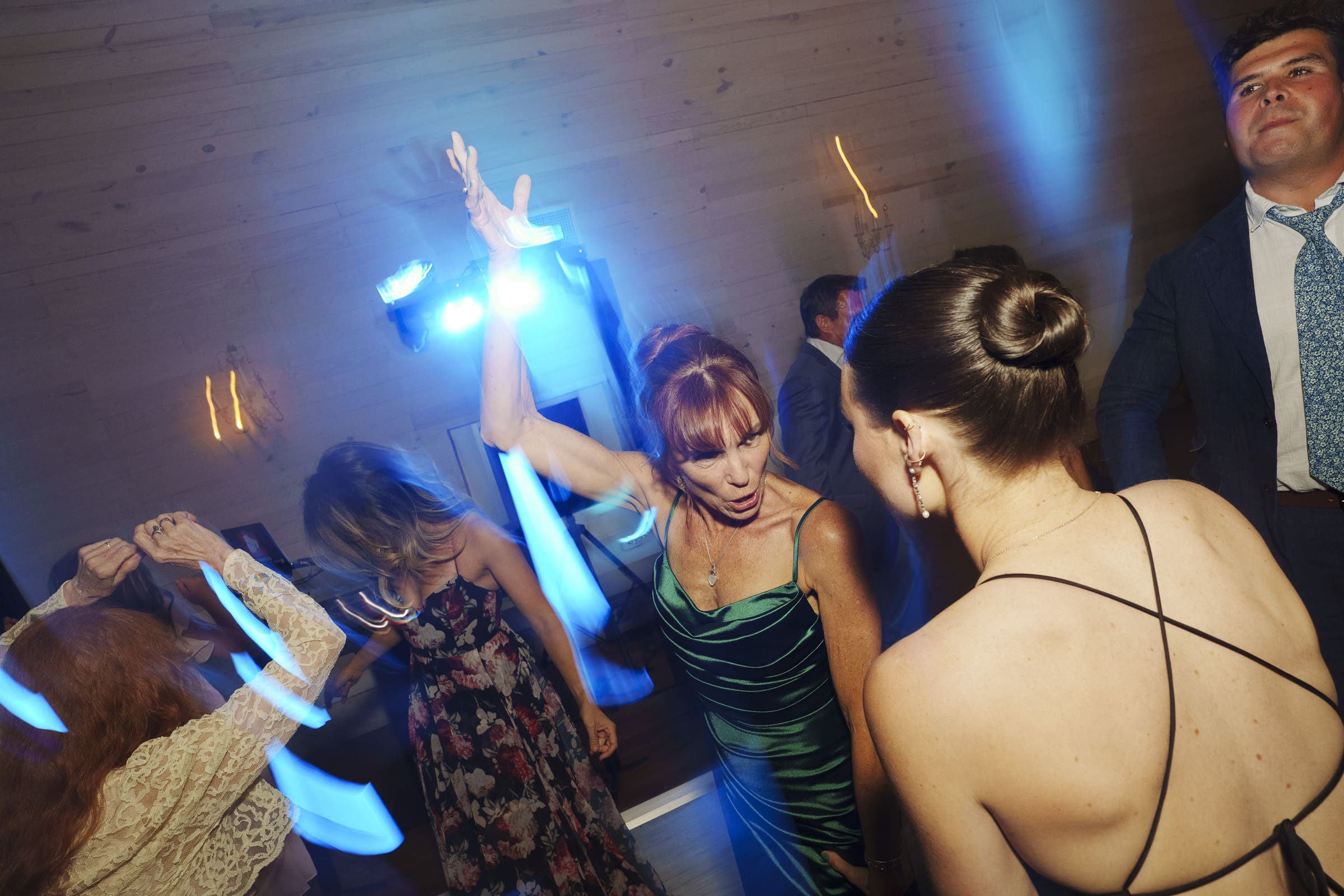 A group of people dressed in formal attire are dancing energetically under blue lights at a Wakefield Barn wedding, with wooden walls in the background. By award-winning Raleigh wedding photographer, Joe Payne.