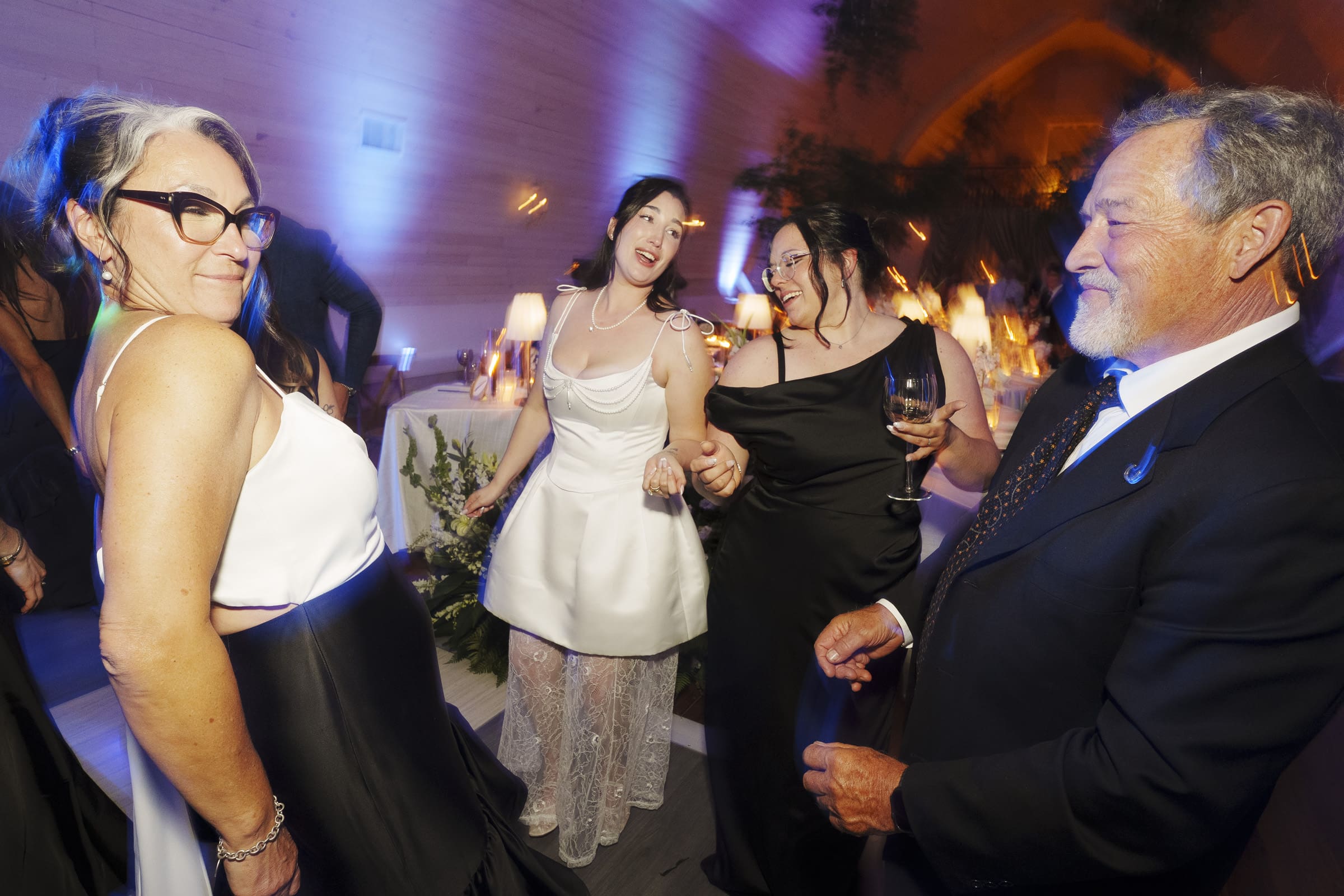 Four people dressed formally, including two women in black dresses, a woman in a white dress, and a man in a suit, smile and dance together at a lively Wakefield Barn wedding with vibrant purple and blue lighting. By award-winning Raleigh wedding photographer, Joe Payne.