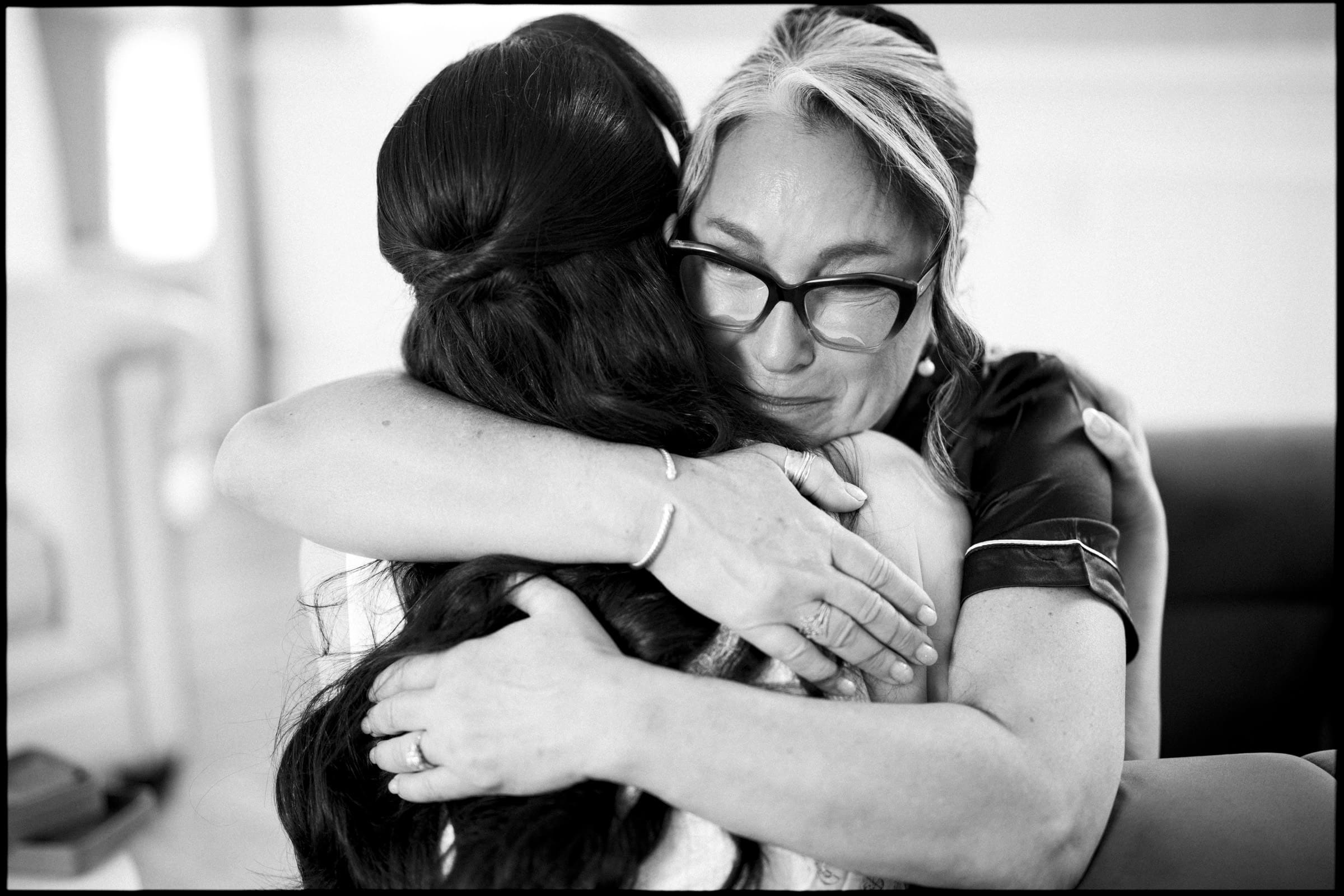 A woman wearing glasses hugs another woman with long dark hair tightly, both appearing emotional and comforted, in a warmly lit indoor setting at a memorable Wakefield Barn wedding. By award-winning Raleigh wedding photographer, Joe Payne.